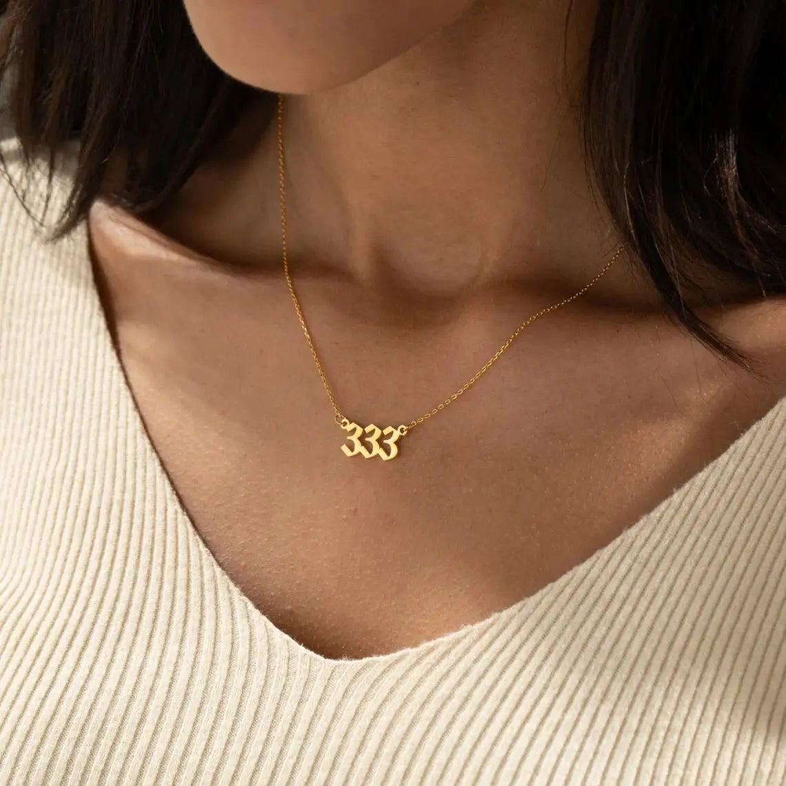 A gold angel number 333 necklace elegantly resting on the collarbone of a woman wearing a beige V-neck top. The pendant shines against her skin, highlighting its elegant design.