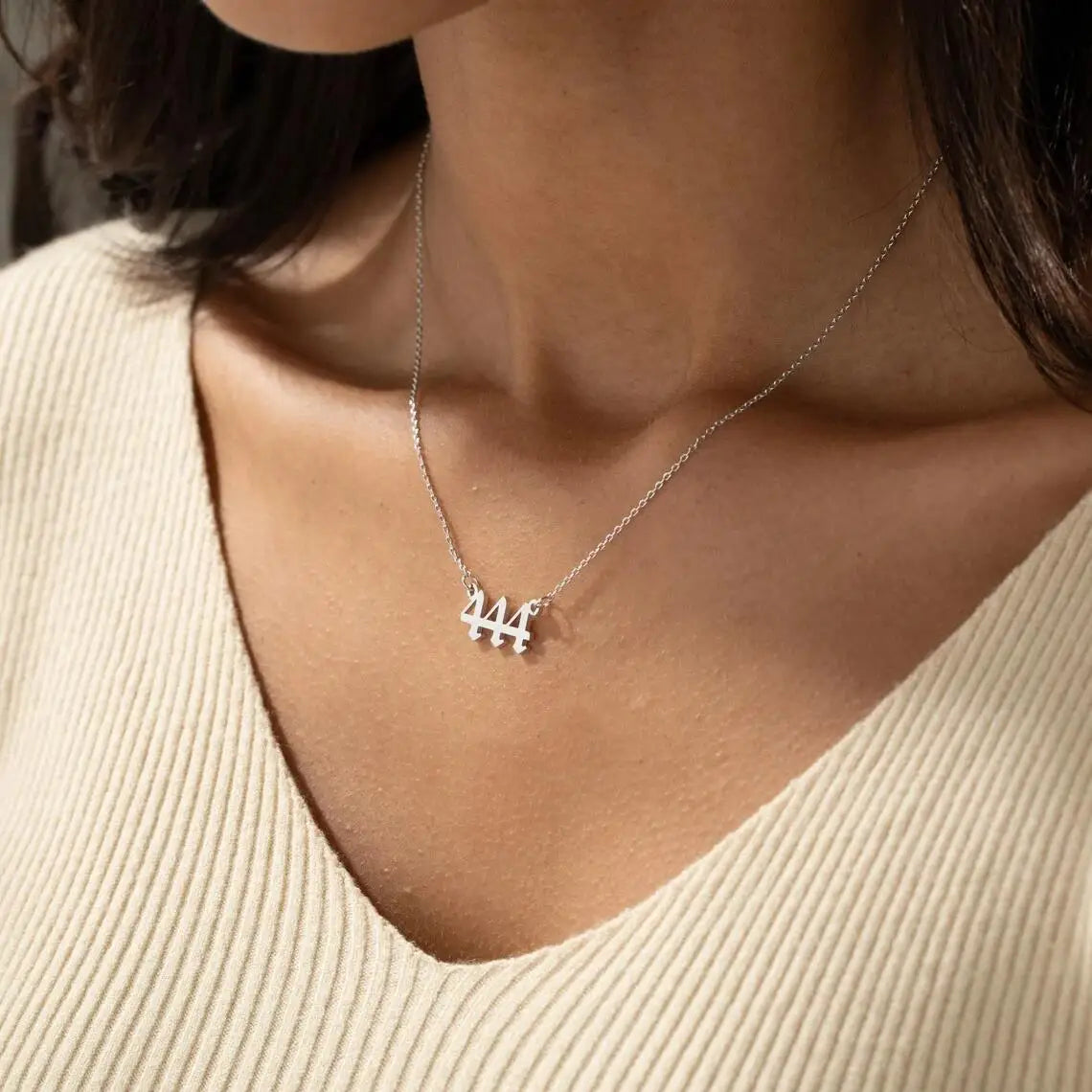 A silver angel number 444 necklace worn by a woman with a delicate chain and a bold, modern number design. The necklace sits elegantly on her collarbone, complementing her beige V-neck top.