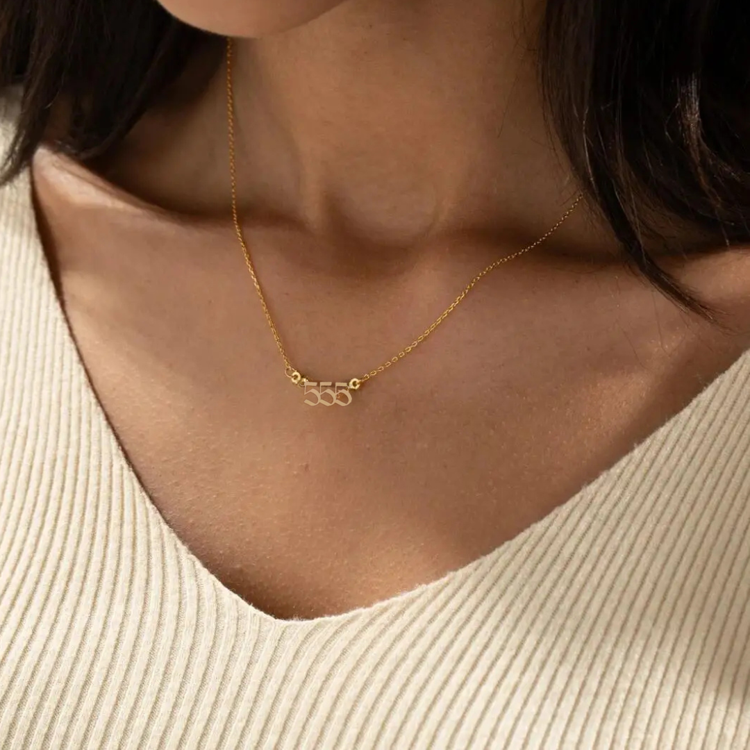 A gold angel number 555 necklace elegantly resting on a woman’s collarbone, complementing her beige knit top. The pendant’s unique design and delicate gold chain add a touch of sophistication.