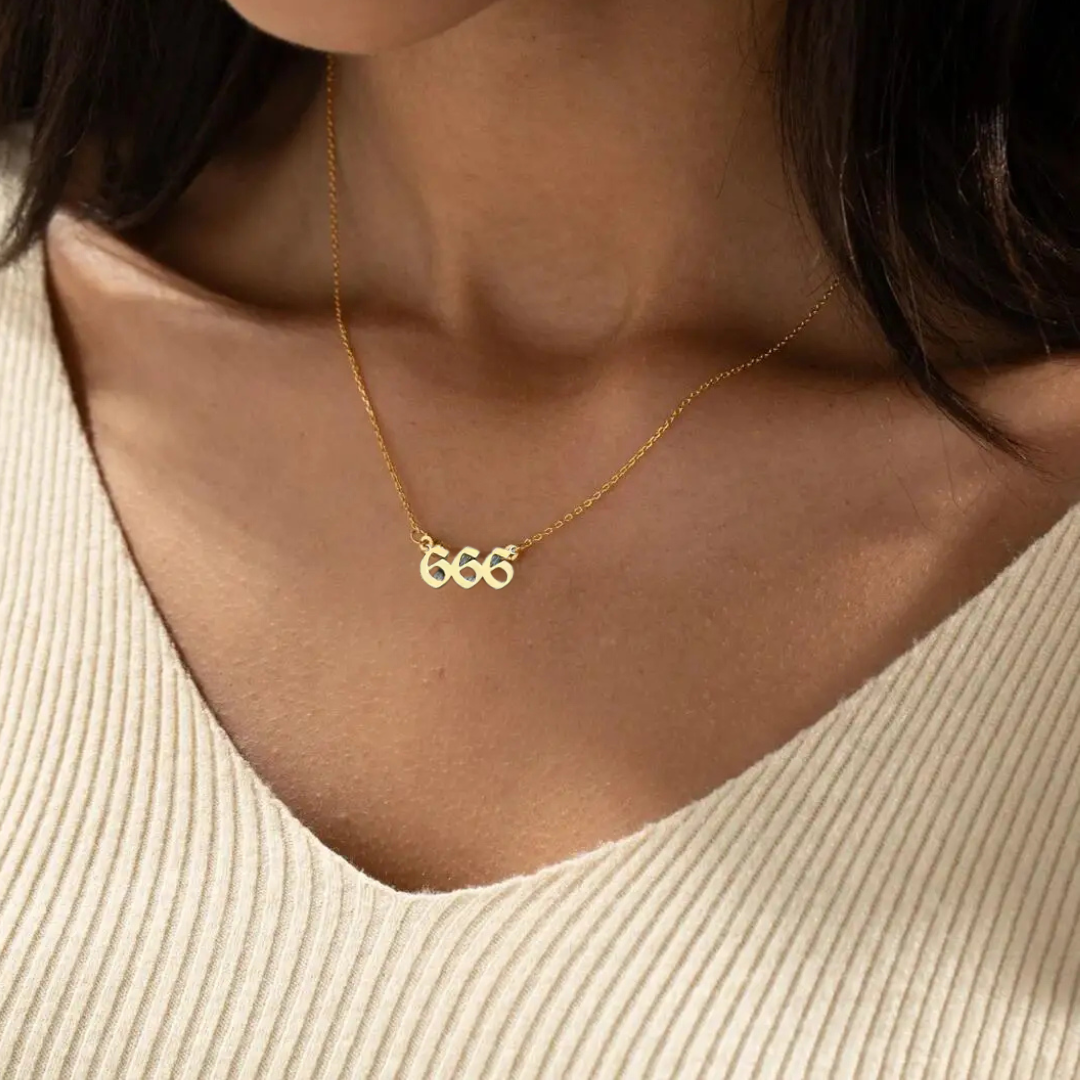 A gold angel number 666 necklace elegantly resting on a woman’s collarbone, complementing her beige knit top. The pendant’s unique design and delicate gold chain add a touch of sophistication.