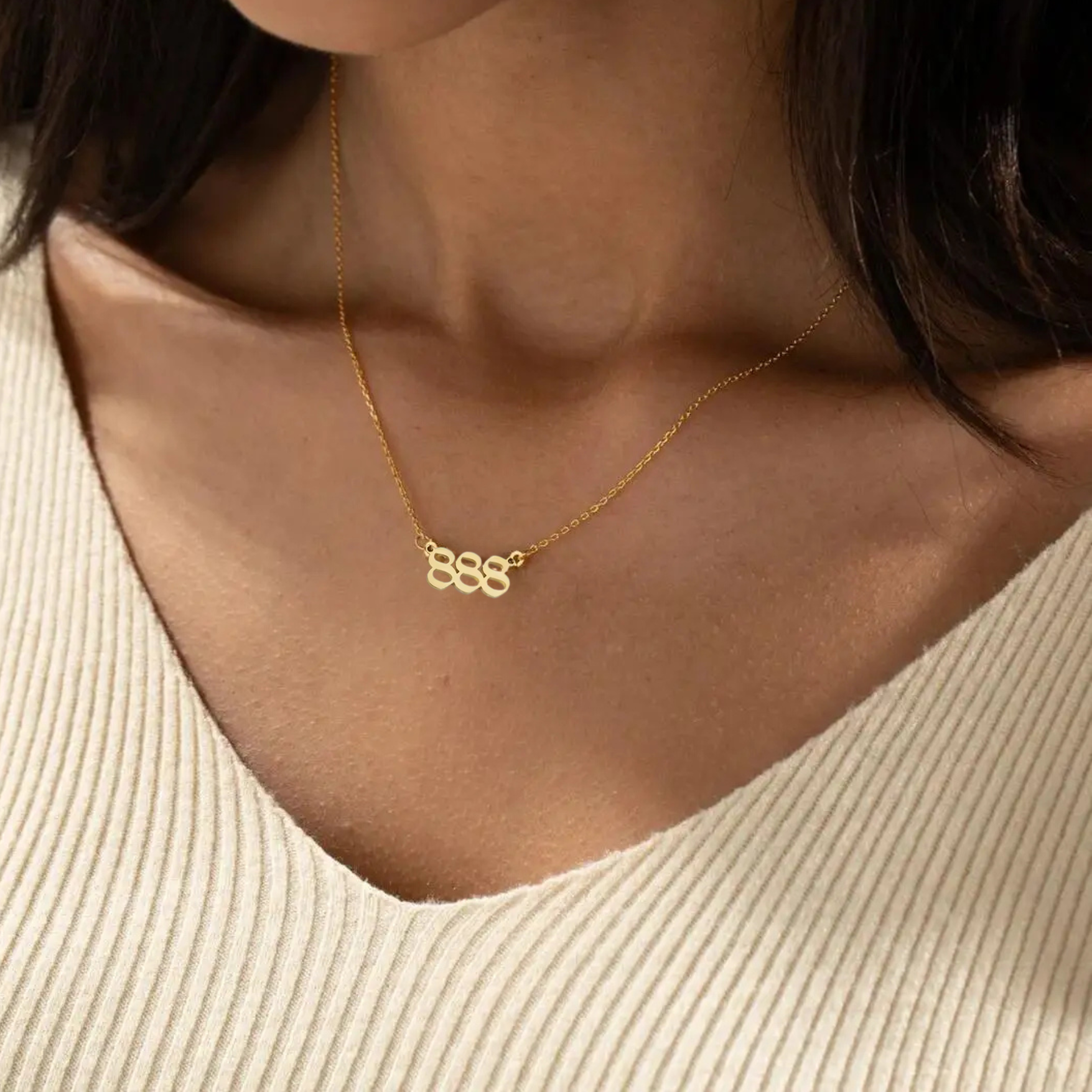 A gold angel number 888 necklace elegantly resting on a woman’s collarbone, complementing her beige knit top. The pendant’s unique design and delicate gold chain add a touch of sophistication.