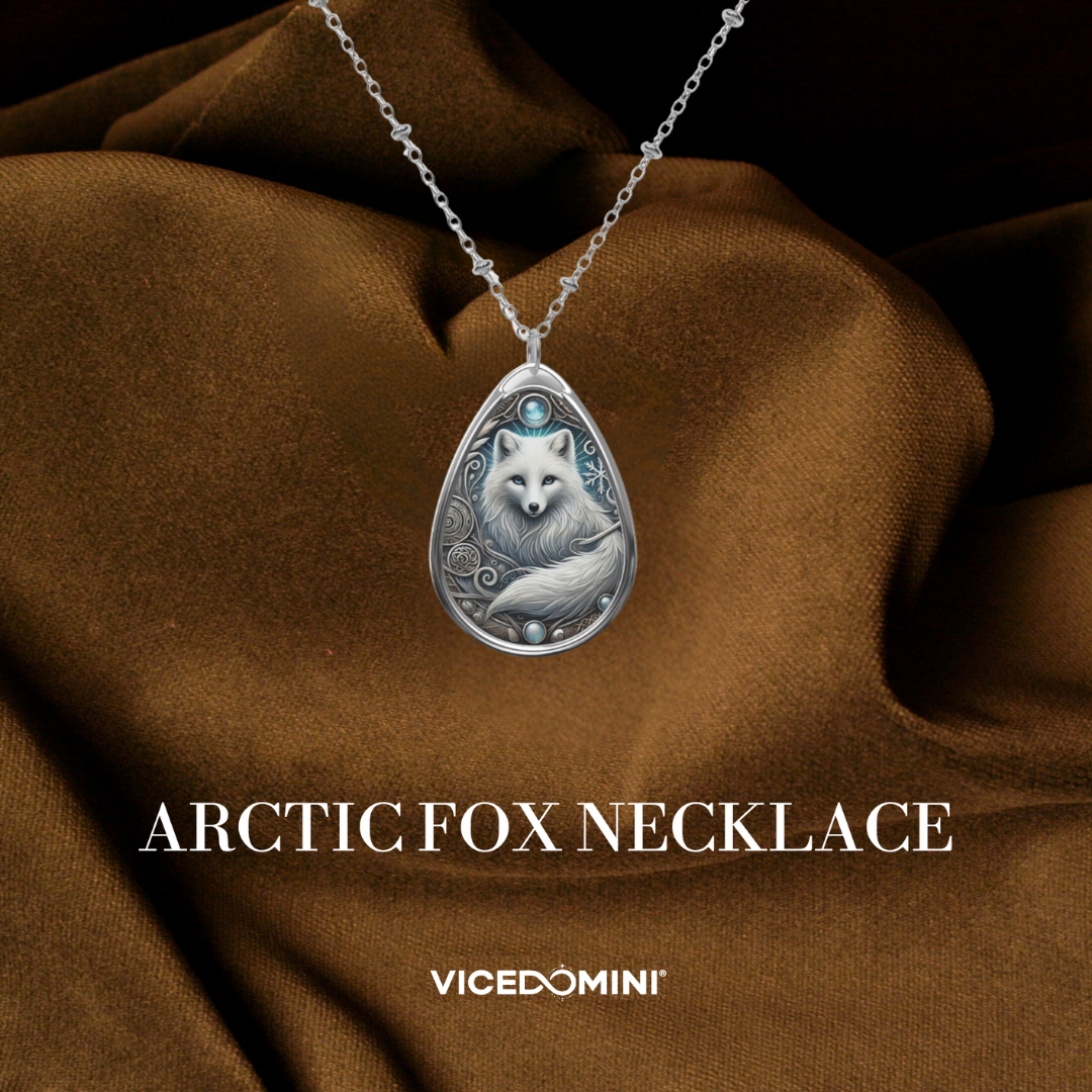 Silver-toned teardrop pendant necklace featuring an engraved Arctic fox design with a small blue gemstone at the top, displayed on draped brown fabric. Text below reads 'ARCTIC FOX NECKLACE' and 'VICEDOMINI'.