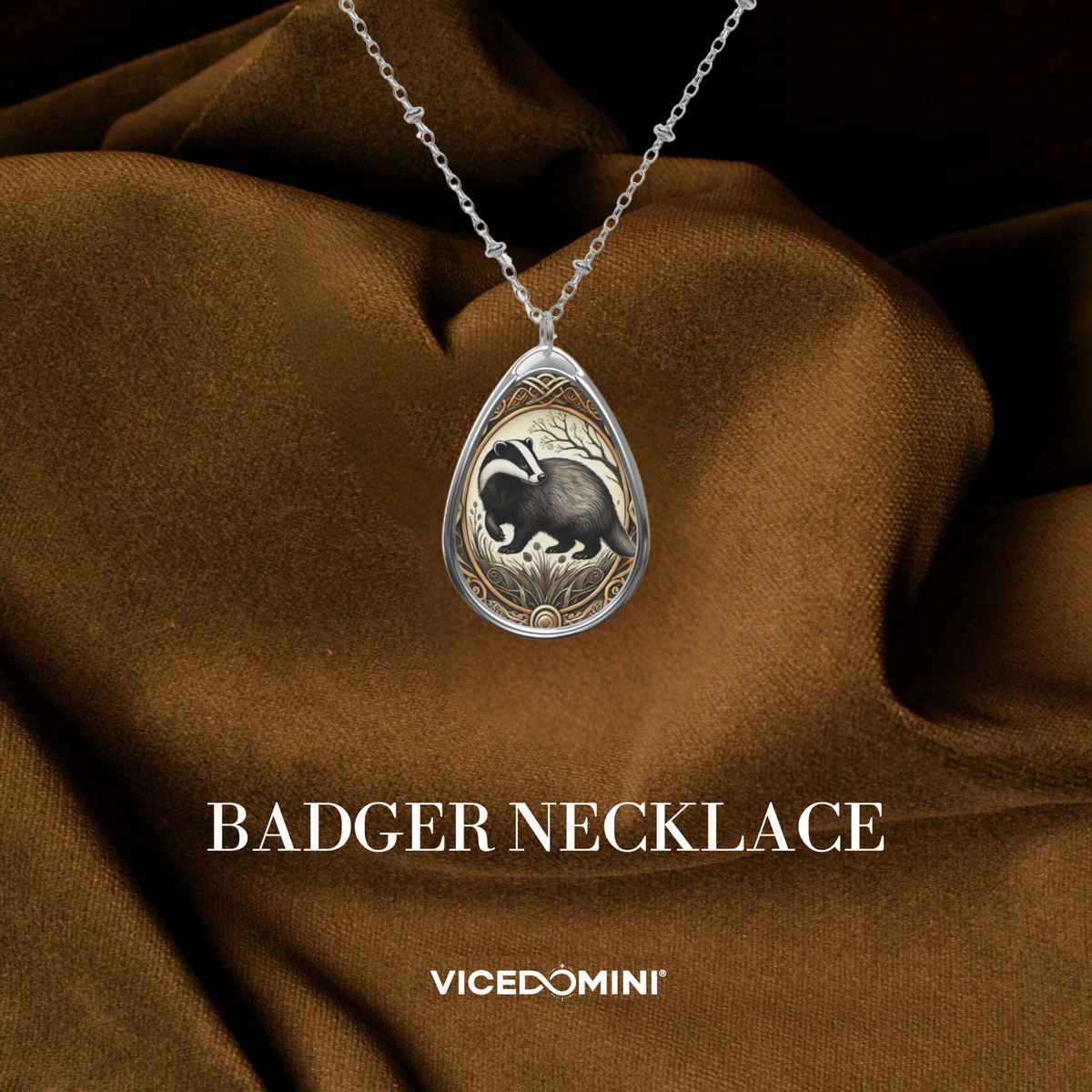 A teardrop-shaped silver pendant necklace featuring an engraved badger design, displayed on draped brown fabric with the text 'BADGER NECKLACE' and 'VICEDOMINI' below.