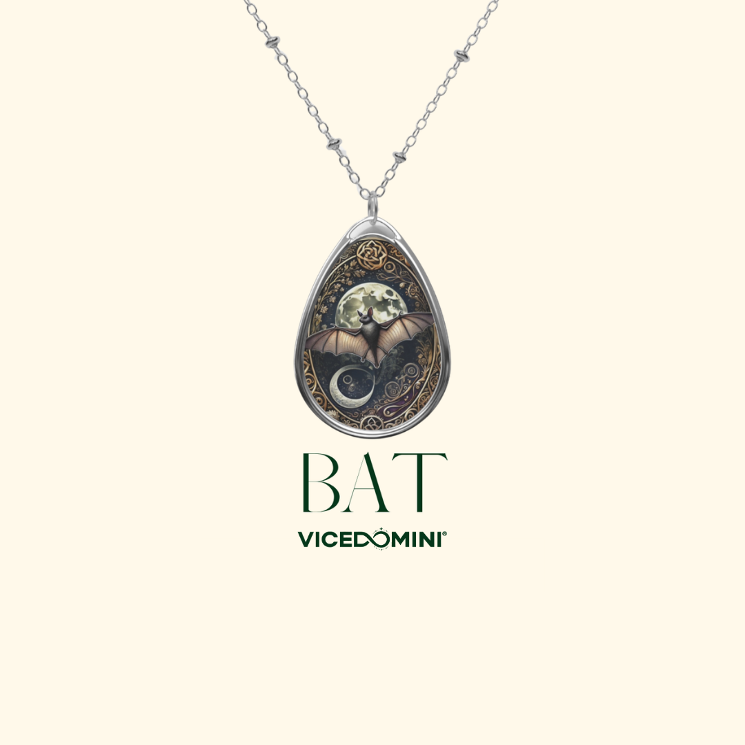 A silver teardrop pendant necklace featuring a detailed bat illustration with a crescent moon and gold decorative accents, shown on a light cream background with the brand text 'BAT VICEDOMINI' below.