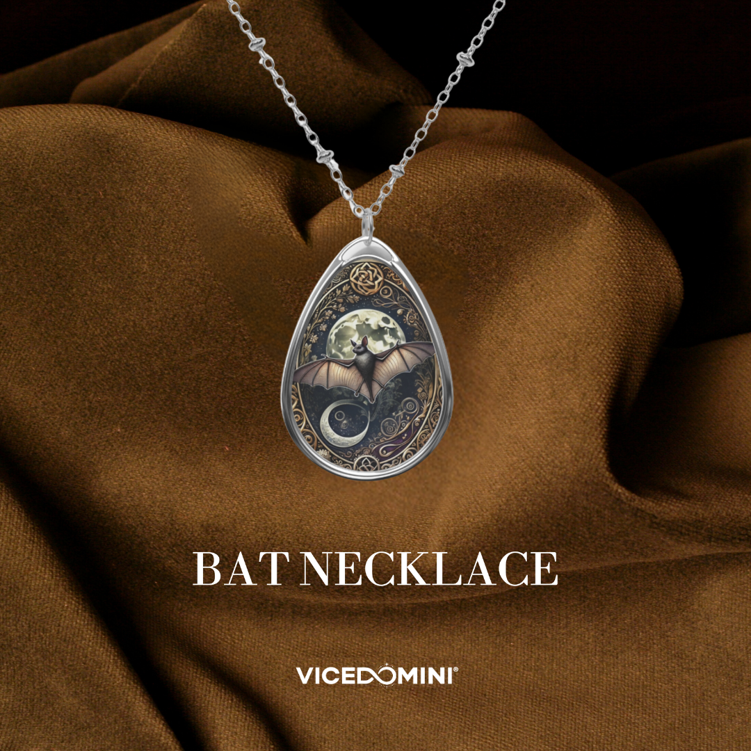 A teardrop-shaped silver pendant necklace featuring a detailed bat illustration with a crescent moon and floral accents, displayed on draped brown fabric with the text 'BAT NECKLACE' and 'VICEDOMINI' below.