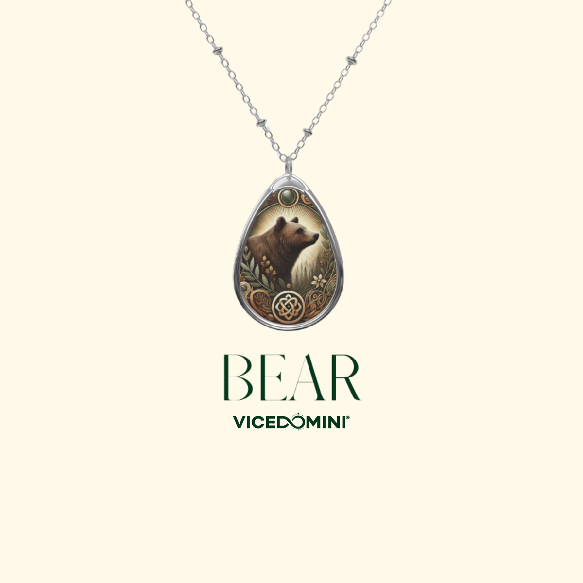 A silver teardrop pendant necklace featuring an illustration of a brown bear surrounded by autumn leaves and a circular medallion, shown on a cream background with the text 'BEAR VICEDOMINI' below.