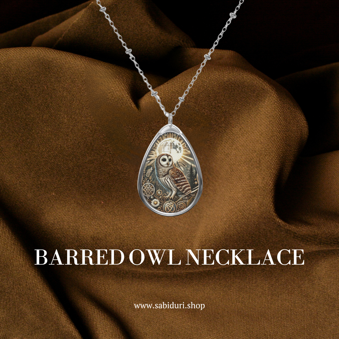A Barred Owl necklace with a silver chain, displayed on a rich brown fabric. The pendant is teardrop-shaped, featuring an intricately designed owl, with the text "BARRED OWL NECKLACE" and the website "www.sabiduri.shop" below it.