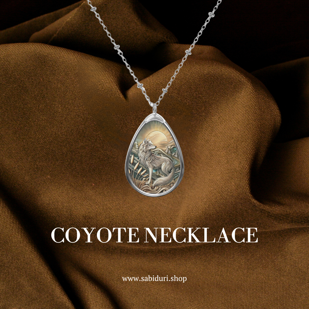 A coyote spirit animal necklace, featuring a teardrop-shaped pendant with an engraved coyote and sunrise scene, is displayed against a rich brown fabric backdrop. The text "COYOTE NECKLACE" and the website www.sabiduri.shop are prominently shown below the pendant.