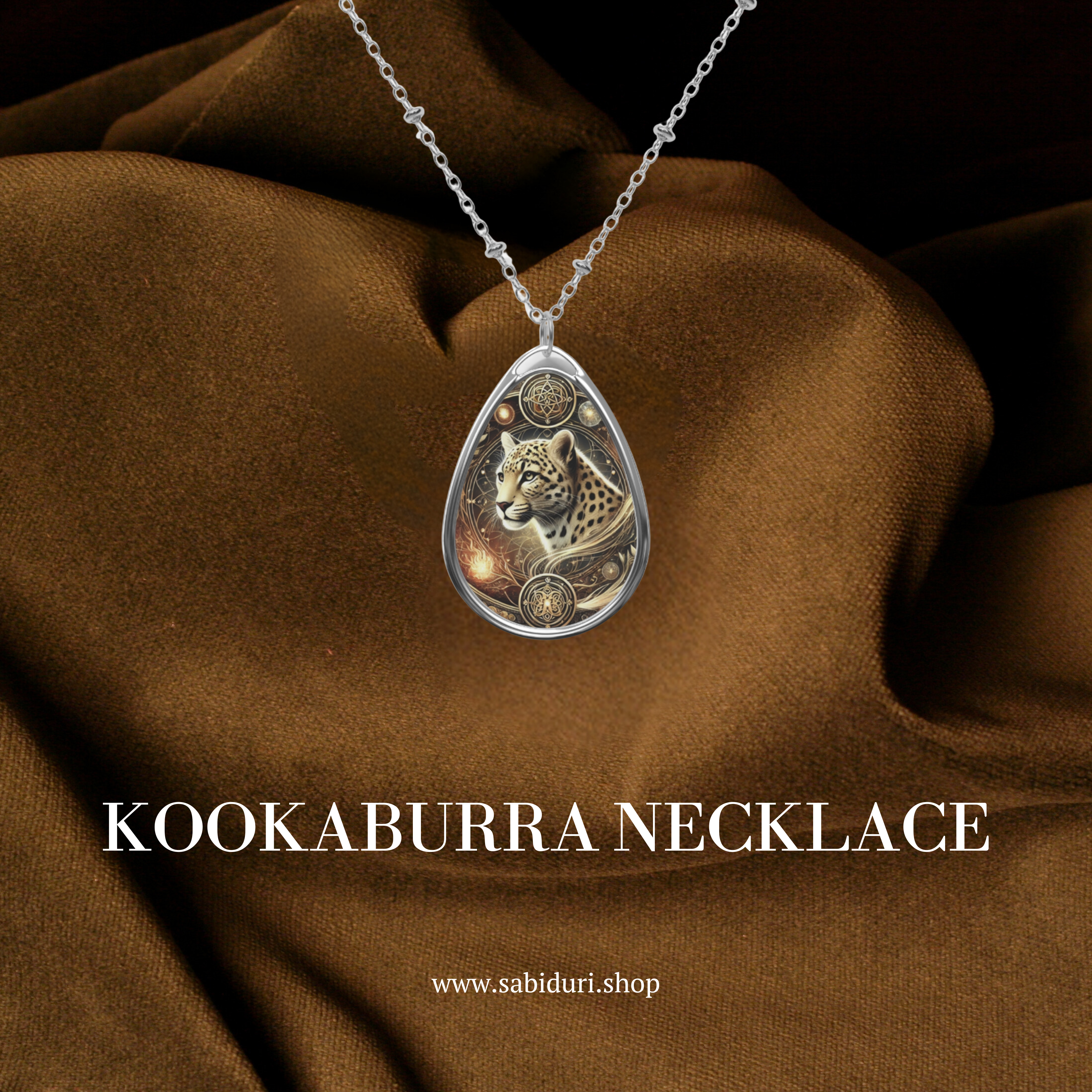 A teardrop-shaped silver pendant featuring an intricately detailed kookaburra perched on a branch, surrounded by golden filigree and blue-green foliage. The pendant hangs from a delicate silver chain, resting against a rich brown fabric backdrop. The text "KOOKABURRA NECKLACE" and "www.sabiduri.shop" is displayed.