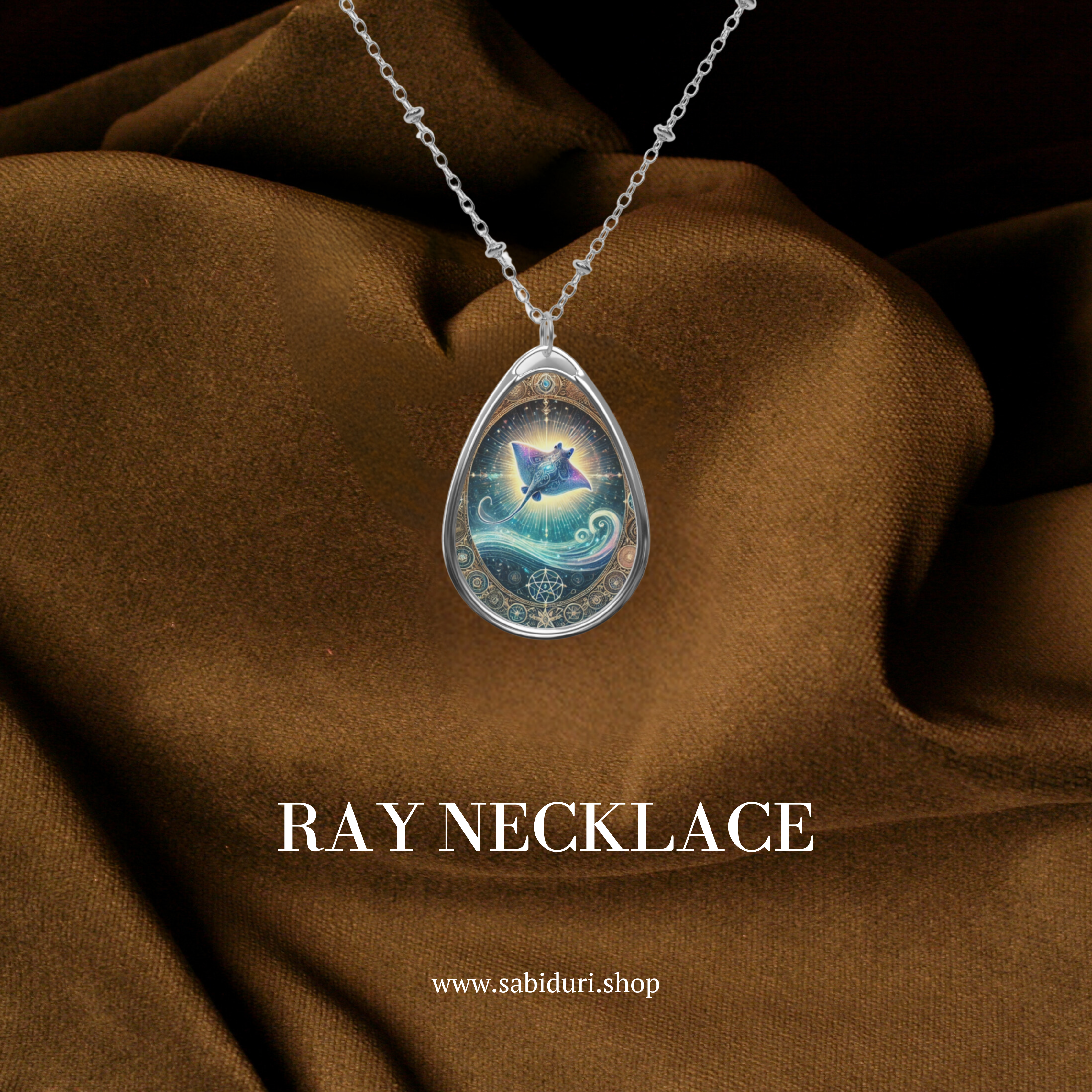 A teardrop-shaped pendant featuring a glowing, celestial-inspired stingray design, surrounded by intricate golden filigree and swirling ocean waves. The pendant hangs from a delicate silver chain, displayed on a luxurious brown fabric background. The text "RAY NECKLACE" and "www.sabiduri.shop" are printed below.