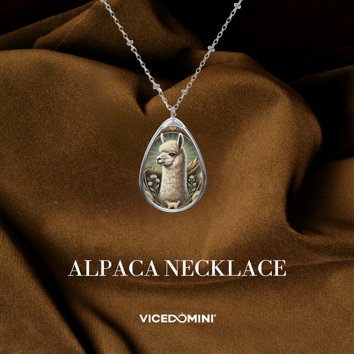 A teardrop-shaped silver pendant necklace featuring an illustrated alpaca, draped over brown satin fabric. The text 'ALPACA NECKLACE' and the brand 'VICEDOMINI' are displayed below.