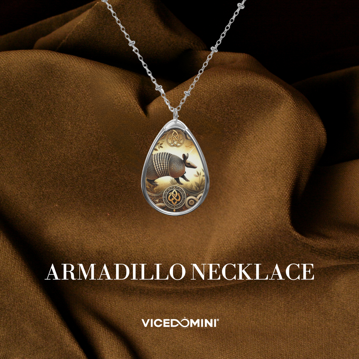 Silver teardrop pendant necklace featuring an intricate armadillo illustration, draped over brown satin fabric with 'ARMADILLO NECKLACE' and 'VICEDOMINI' text below.