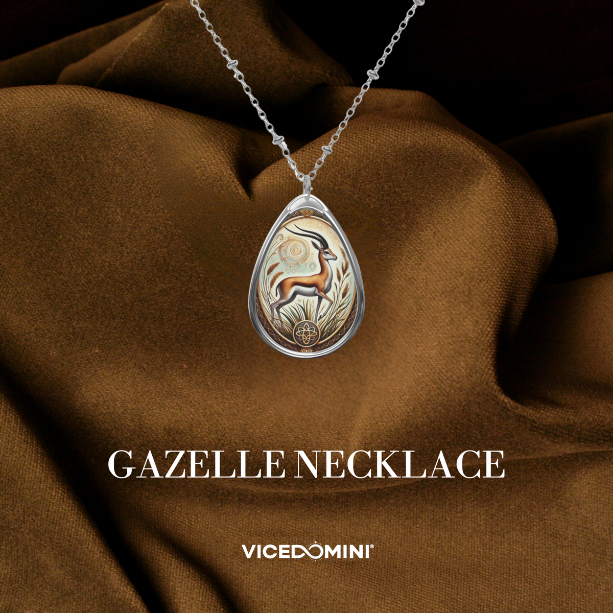 A teardrop-shaped silver pendant necklace featuring an engraved gazelle motif on a warm metallic background, draped elegantly over brown satin fabric. The text 'GAZELLE NECKLACE' and 'VICEDOMINI' are displayed below.