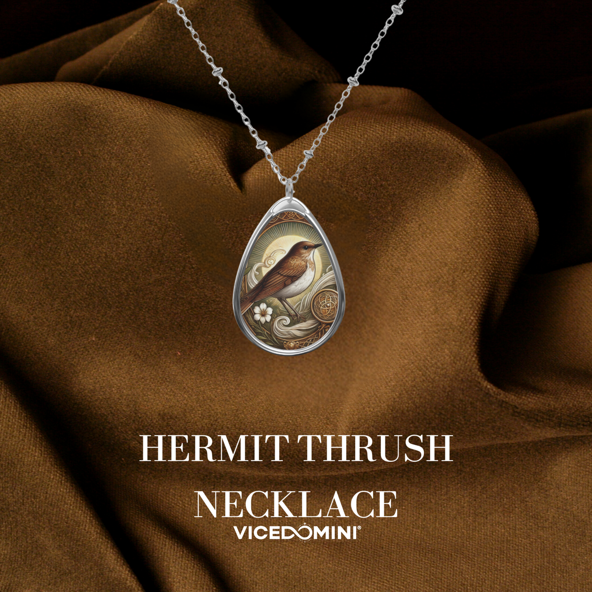 A teardrop-shaped pendant necklace featuring an illustrated hermit thrush bird in a silver frame, draped over brown satin fabric with the text 'HERMIT THRUSH NECKLACE' and 'VICEDOMINI' below.