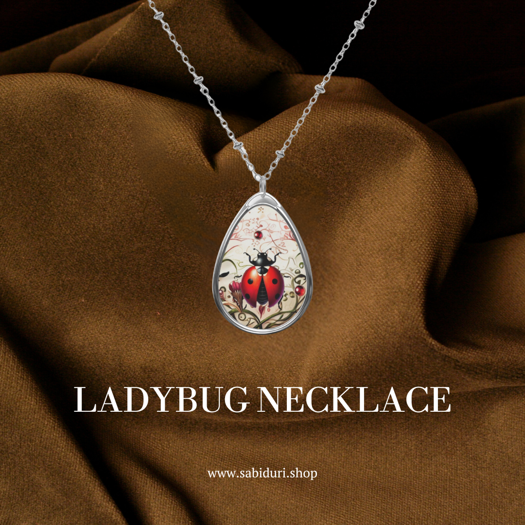 A silver necklace with a teardrop-shaped pendant showcasing a red ladybug illustration, placed against a rich brown fabric backdrop. The text "LADYBUG NECKLACE" and "www.sabiduri.shop" are displayed prominently in white at the bottom.