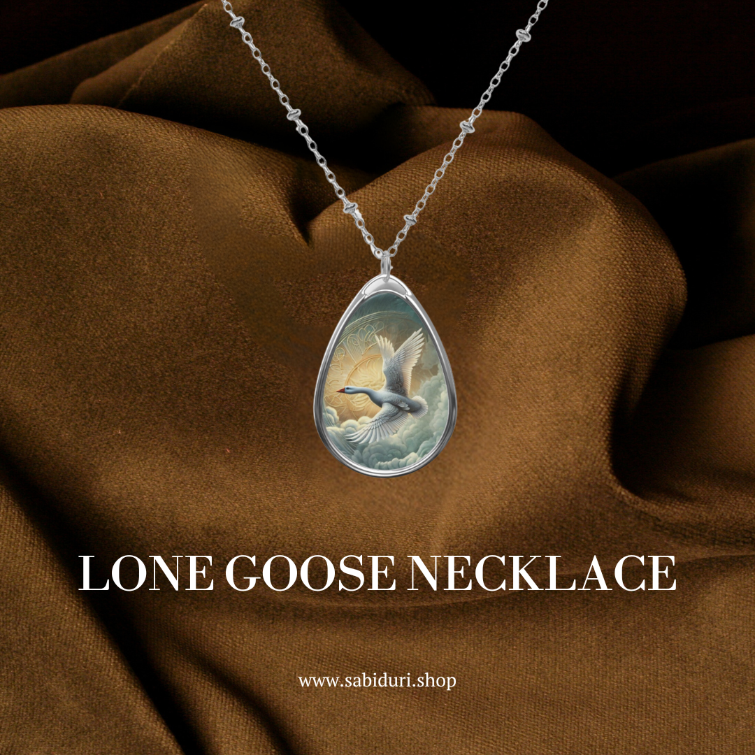A teardrop-shaped necklace pendant showcasing a painting of a goose soaring in front of a glowing sun, set against a rich brown fabric. The image includes the text "Lone Goose Necklace" and "www.sabiduri.shop," emphasizing the product and its brand.