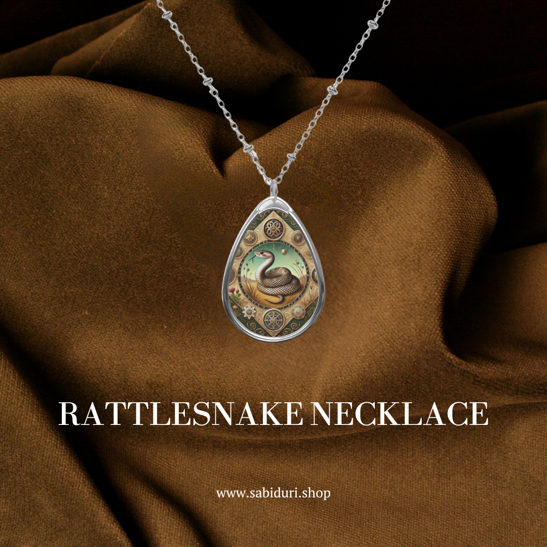 A teardrop-shaped pendant with a coiled rattlesnake illustration, surrounded by gears and floral motifs, hanging against a backdrop of rich brown fabric. The text "Rattlesnake Necklace" and "www.sabiduri.shop" is displayed at the bottom.