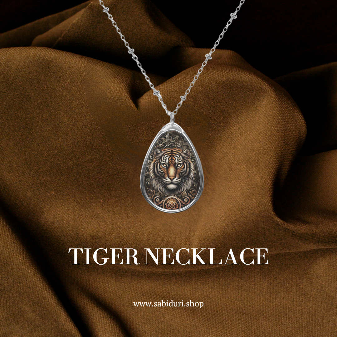 A silver necklace with a teardrop-shaped pendant featuring an intricate tiger design, displayed on a rich brown fabric background. The text "TIGER NECKLACE" is positioned below the pendant, along with the website URL "www.sabiduri.shop."