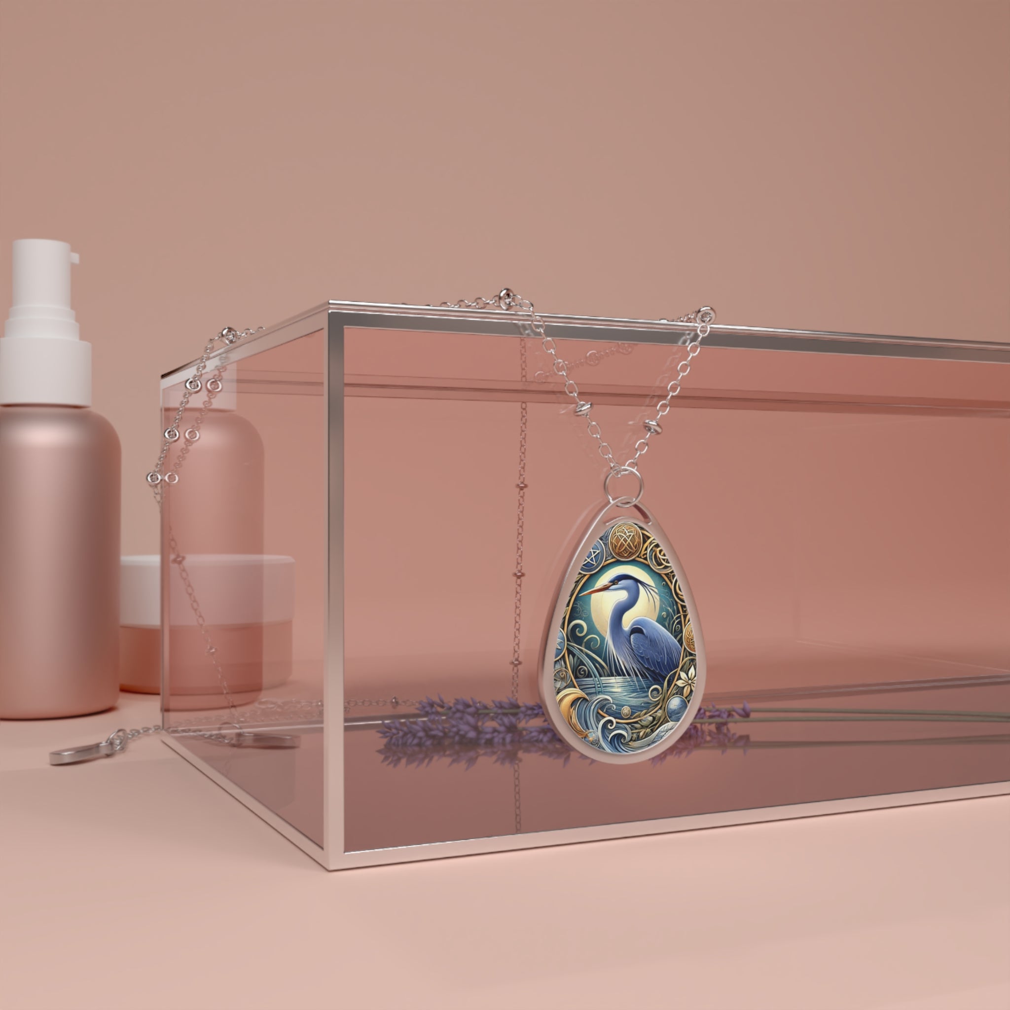 A blue heron pendant necklace draped over a clear box with lavender and cosmetics, set against a soft pink background. The pendant depicts a calm blue heron within a detailed natural landscape, symbolizing grace and self-reflection.