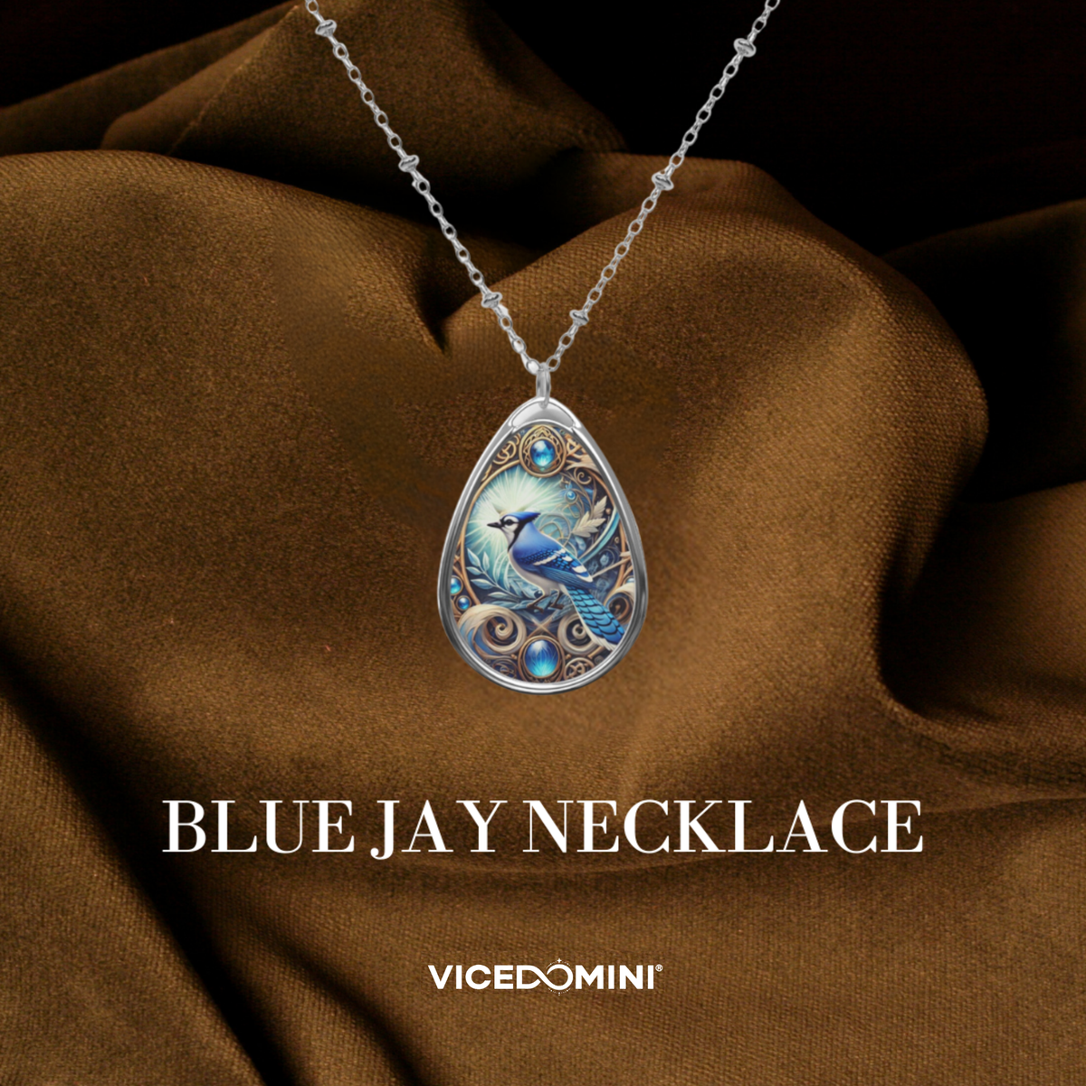 A teardrop-shaped pendant necklace with a detailed blue bird illustration on a silver-toned chain, draped over brown satin fabric. The text 'BLUE JAY NECKLACE' and the brand 'VICEDOMINI' are displayed below.