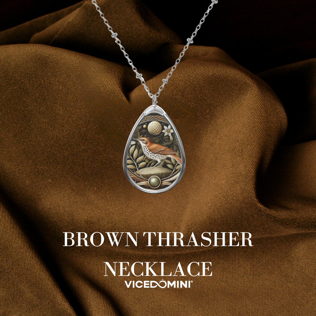 A teardrop-shaped silver pendant necklace featuring an intricate illustration of a brown thrasher bird, set against a background of draped brown satin fabric. The text 'BROWN THRASHER NECKLACE' and the brand 'VICEDOMINI' are displayed below the necklace.