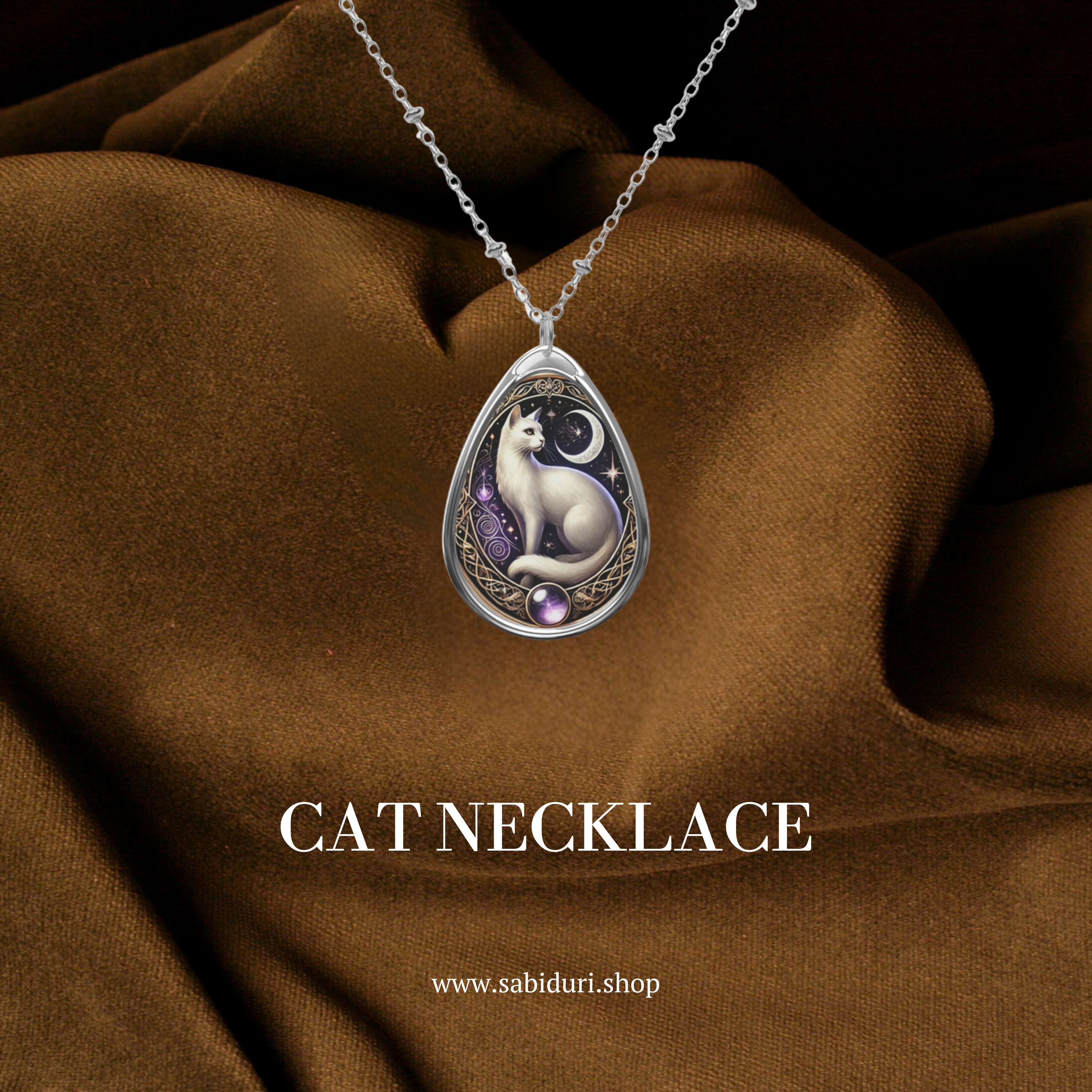 A silver cat-themed pendant with a white feline design, moonlit background, and ornate golden embellishments, displayed on rich brown fabric. The text "CAT NECKLACE" and "www.sabiduri.shop" are elegantly placed below.