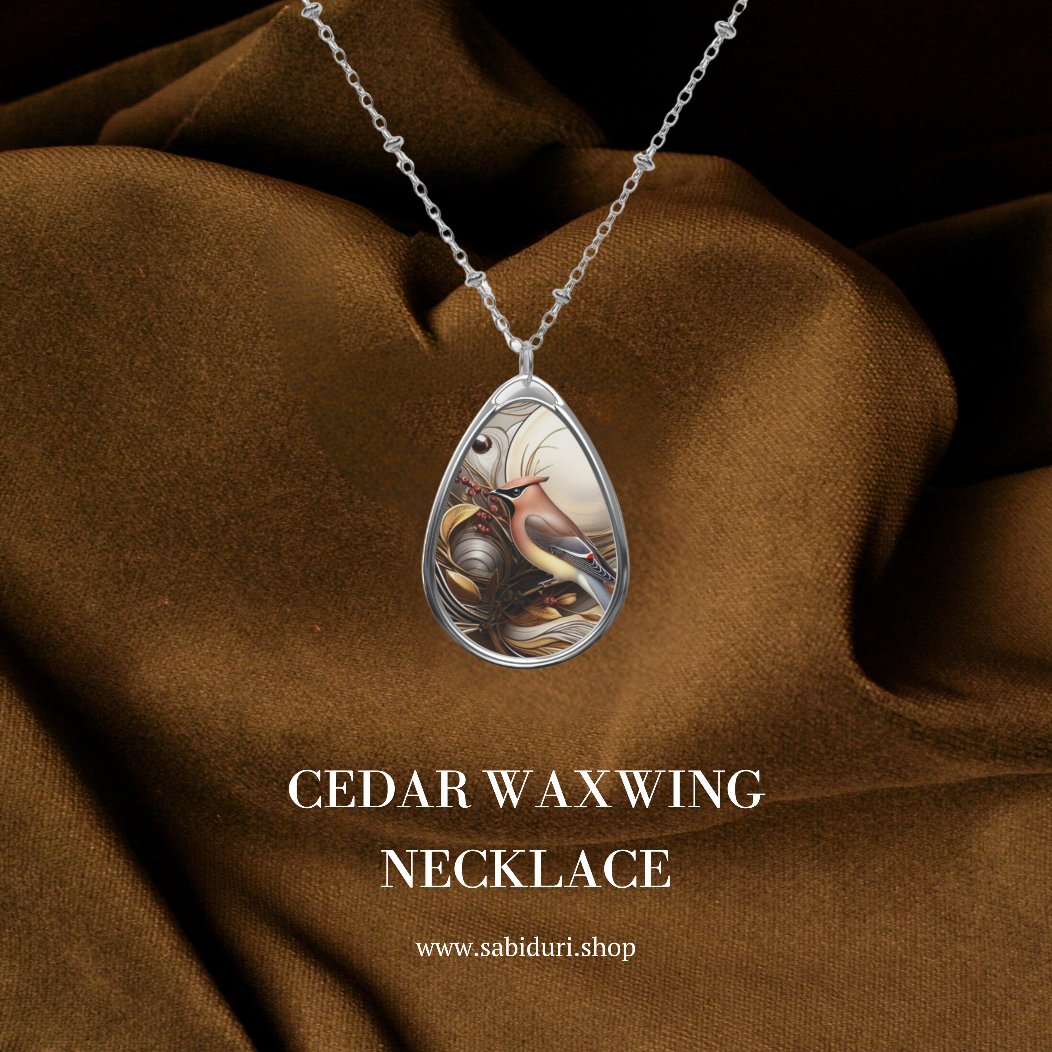 A close-up of the Cedar Waxwing Spirit Animal necklace with a teardrop-shaped pendant featuring an artistic depiction of a cedar waxwing bird. The necklace is draped against a rich brown fabric background, creating a luxurious contrast. The text "Cedar Waxwing Necklace" appears below the pendant, along with the website URL "www.sabiduri.shop."