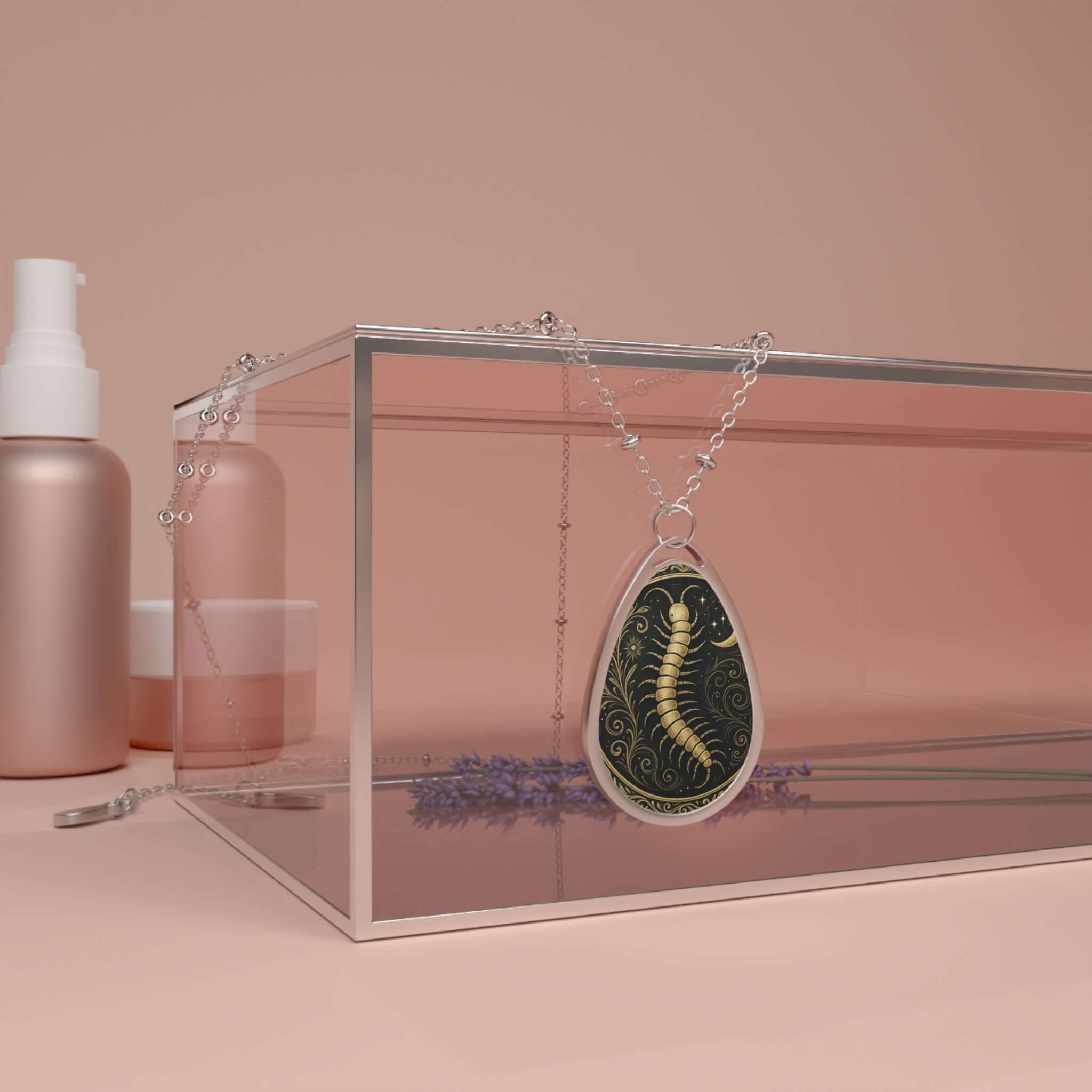 Teardrop-shaped necklace with a gold centipede design set against a swirling black background, draped over a pink glass box beside lavender and skincare items.
