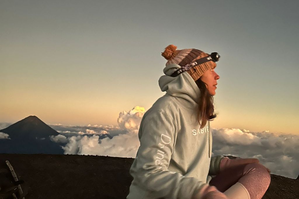A person in a cozy hat and Sabiduri hoodie sits contemplatively atop a mountain at sunrise, with the silhouette of a neighboring peak visible against a soft sky of clouds.