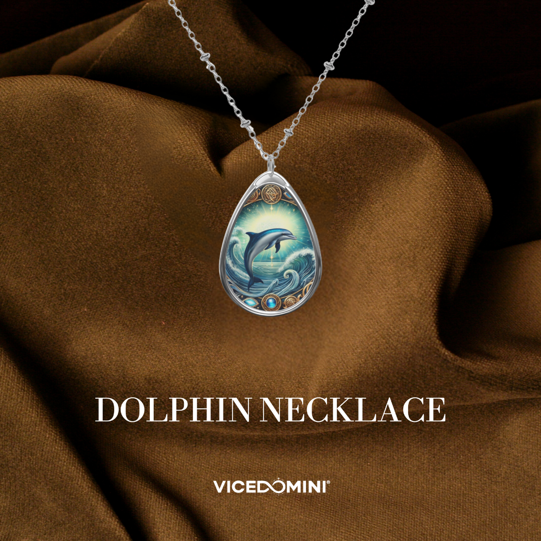 A teardrop-shaped silver pendant necklace featuring an intricate blue and green dolphin and wave design, displayed on draped brown fabric with the text 'DOLPHIN NECKLACE' and 'VICEDOMINI' below.