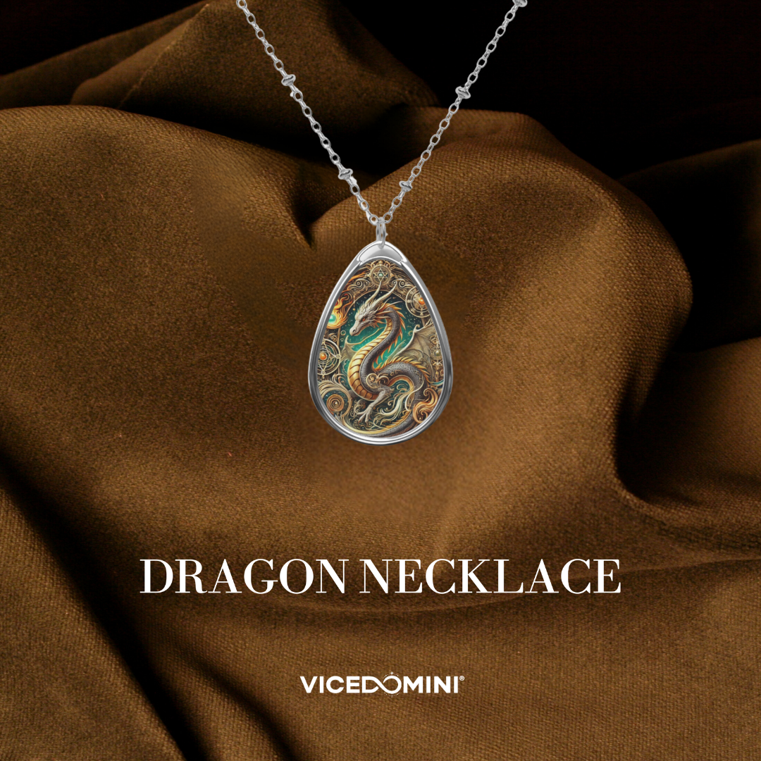 A teardrop-shaped silver pendant necklace featuring an intricate, colorful dragon illustration, displayed on draped brown satin fabric with the text 'DRAGON NECKLACE' and 'VICEDOMINI' below.