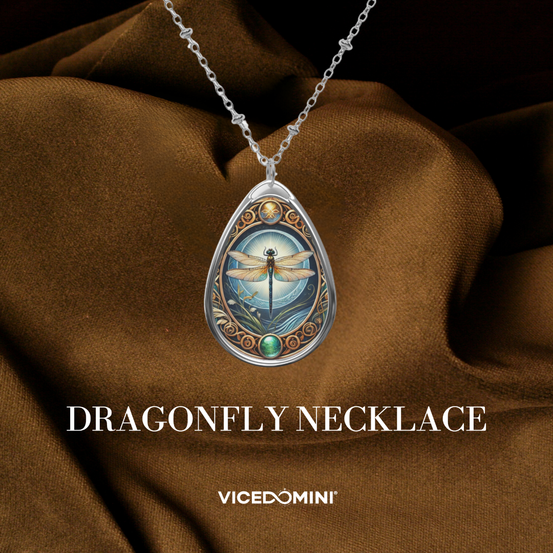 A teardrop-shaped silver pendant necklace featuring an intricate dragonfly illustration with blue, gold, and green details, displayed on draped brown fabric. The text 'DRAGONFLY NECKLACE' and the brand 'VICEDOMINI' are visible below.