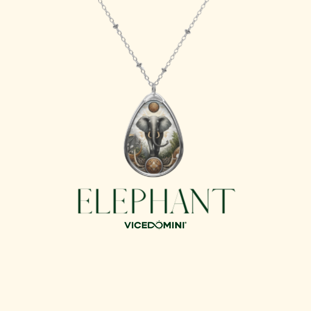 A teardrop-shaped pendant necklace featuring an elephant illustration with green foliage and gold-toned accents, suspended from a silver chain on a cream background. The text 'ELEPHANT' and 'VICEDOMINI' appears below the pendant.