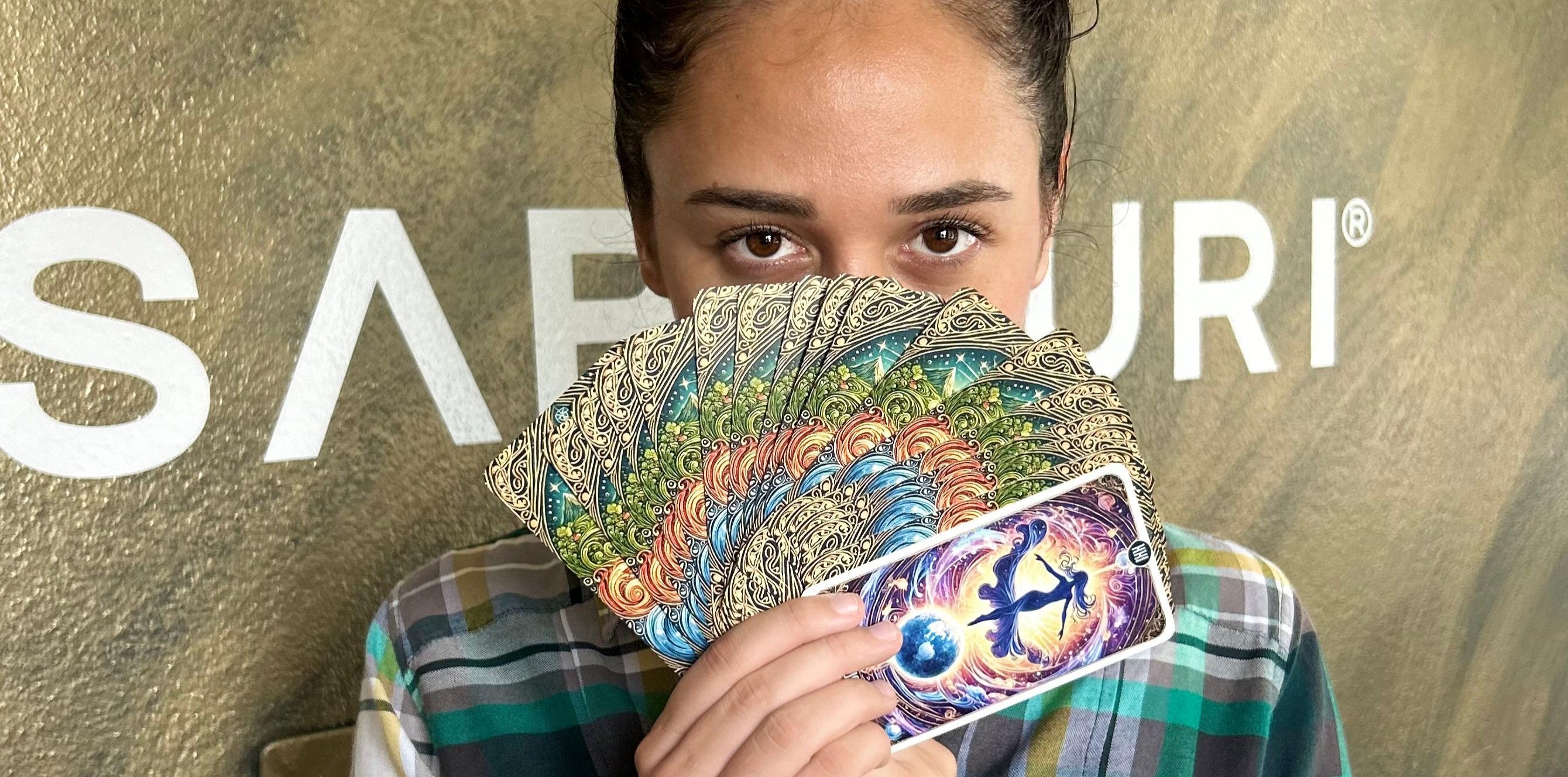 A person holds a fan of intricately designed tarot cards, mostly concealing their face except for their eyes. The tarot cards feature vibrant colors and patterns, with the card closest to the camera displaying an illustration of a cosmic figure dancing above a glowing Earth. The background shows part of a sign with the letters "SABIDURI" visible.