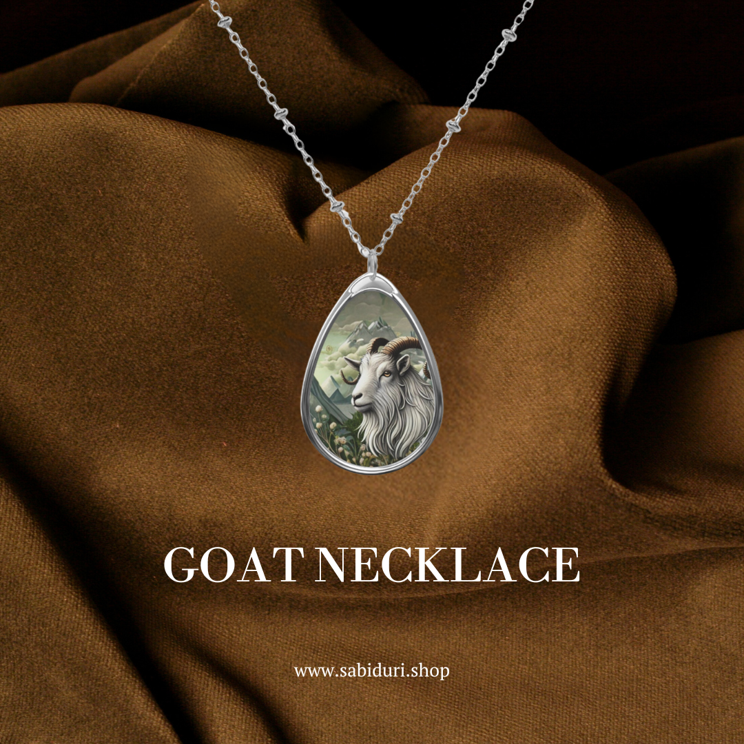A silver goat spirit animal necklace, symbolizing determination and confidence, hanging elegantly against a rich brown fabric background, accompanied by the text "GOAT NECKLACE" and the website "www.sabiduri.shop."
