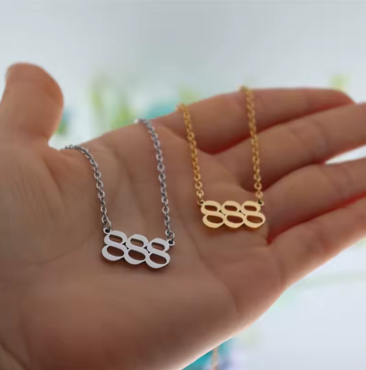 A close-up of a hand holding two angel number 888 necklaces—one in gold and one in silver. The modern pendant design and polished finish reflect light, emphasizing their elegant style.