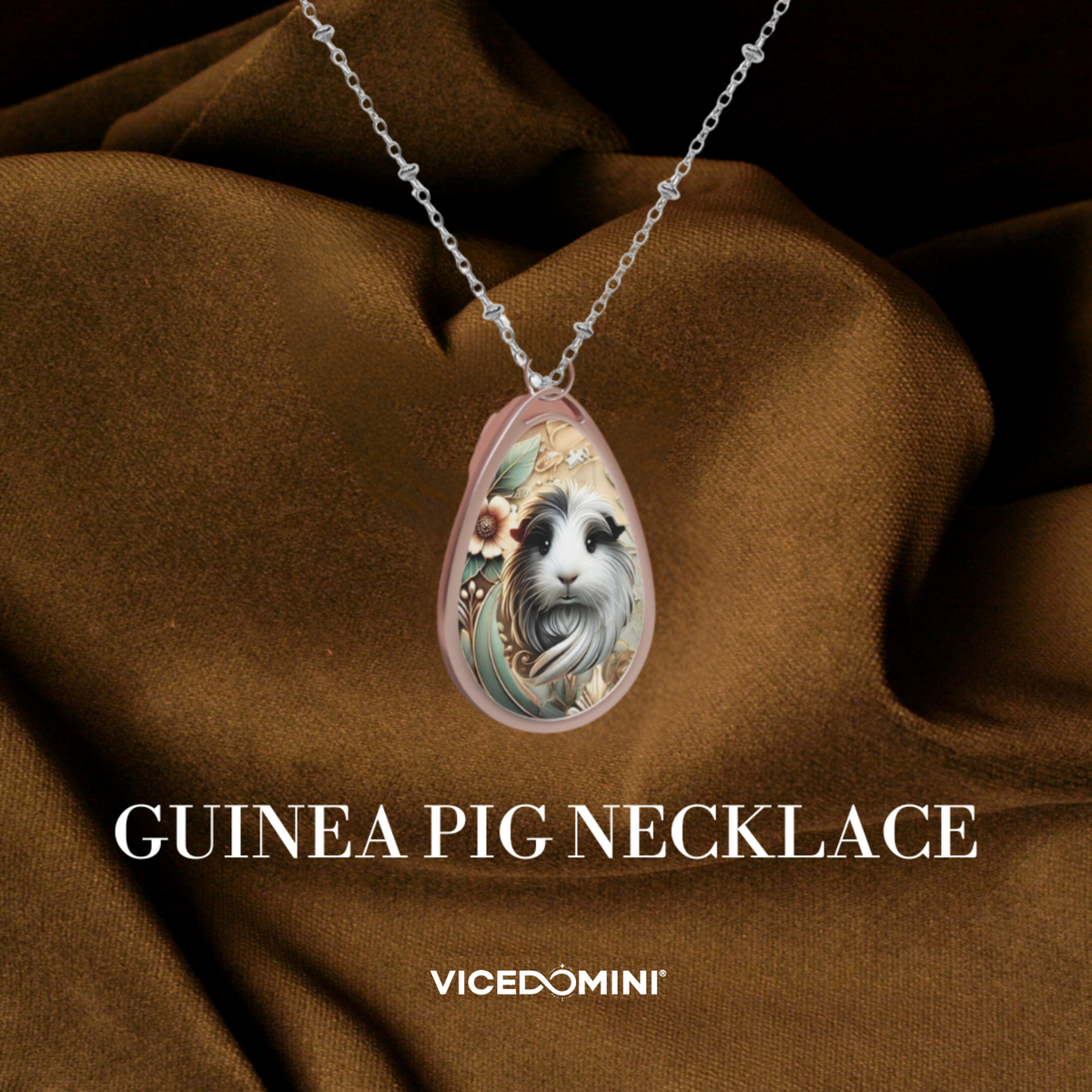 A teardrop-shaped pendant necklace featuring a detailed illustration of a guinea pig, displayed on draped brown fabric. The necklace has a silver chain and a pinkish-beige pendant frame.