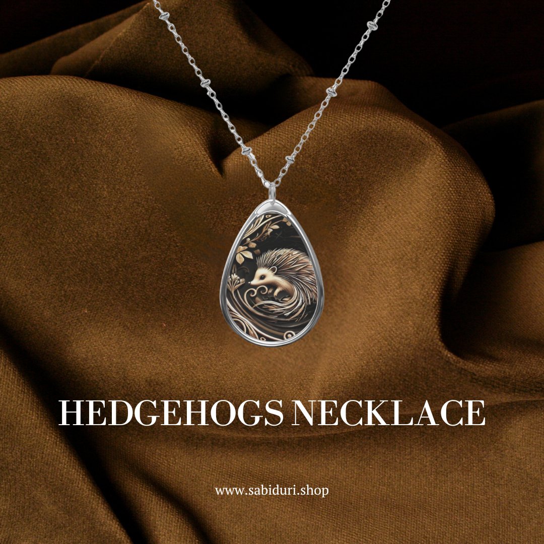 A teardrop-shaped necklace pendant with a hedgehog design, laying against a luxurious, brown fabric backdrop. The text at the bottom reads "Hedgehogs Necklace" along with the website URL "www.sabiduri.shop" in white font.