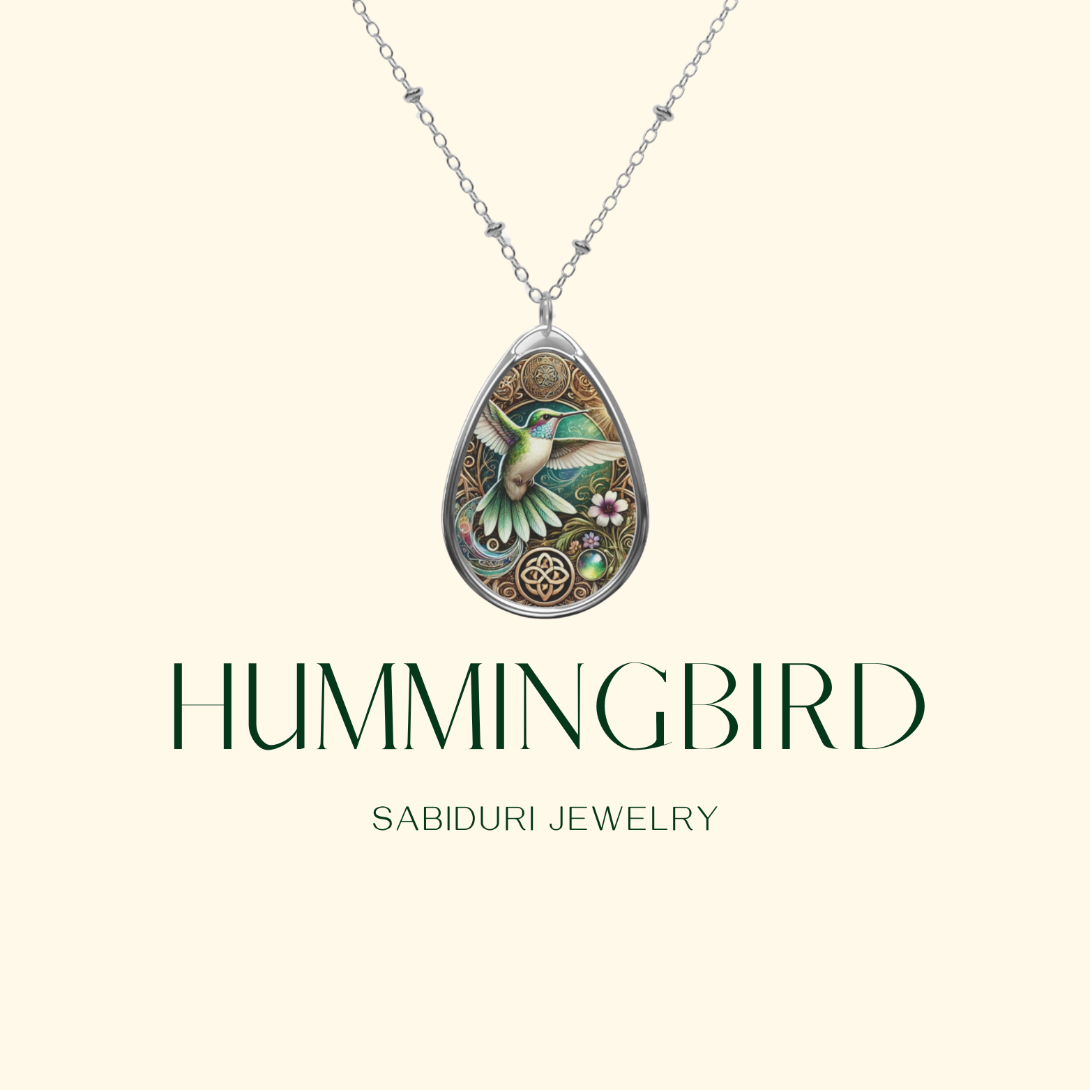 A delicate silver necklace with a teardrop-shaped pendant featuring an intricately designed hummingbird in flight. The bird is surrounded by floral and geometric elements in gold and green tones, evoking a sense of energy and vitality. Below the pendant, "HUMMINGBIRD" and "SABIDURI JEWELRY" are elegantly written.