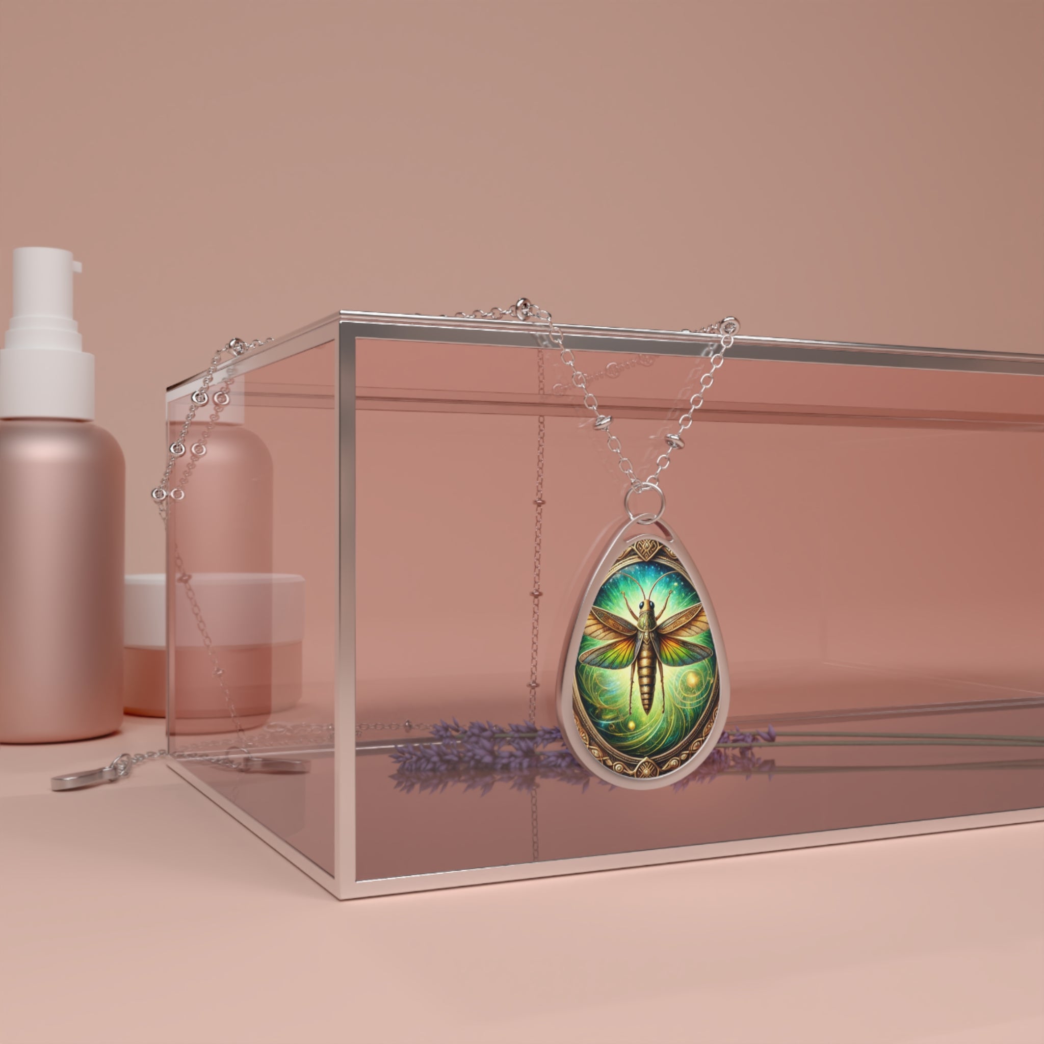 A teardrop-shaped pendant featuring an intricately designed grasshopper with glowing green and gold wings, set against a swirling, nature-inspired background. The pendant hangs from a delicate silver chain, draped over a modern glass display box with soft lavender flowers beneath it. The scene is set against a warm, neutral-toned backdrop.