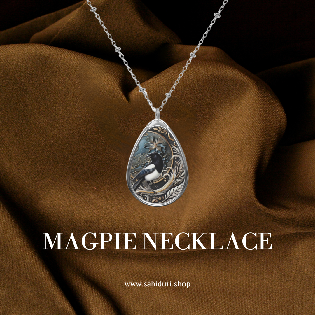 A close-up of the magpie necklace, featuring the same detailed pendant design, resting against a soft brown fabric background. The text "MAGPIE NECKLACE" is written in bold white letters, and below it is the URL "www.sabiduri.shop."