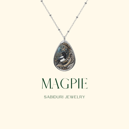 A close-up of a teardrop-shaped pendant featuring a detailed illustration of a magpie with intricate gold and silver accents. The necklace is displayed on a simple light background with the text "MAGPIE" and "SABIDURI JEWELRY" beneath it.