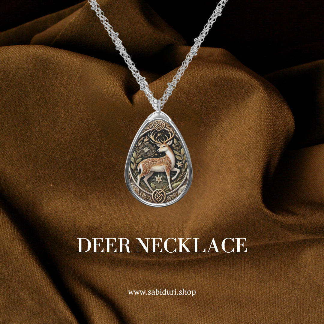 A close-up of a silver teardrop pendant showcasing an intricate deer design surrounded by ornamental patterns, displayed against a rich brown fabric background. Text on the image reads "DEER NECKLACE" and the website "www.sabiduri.shop.