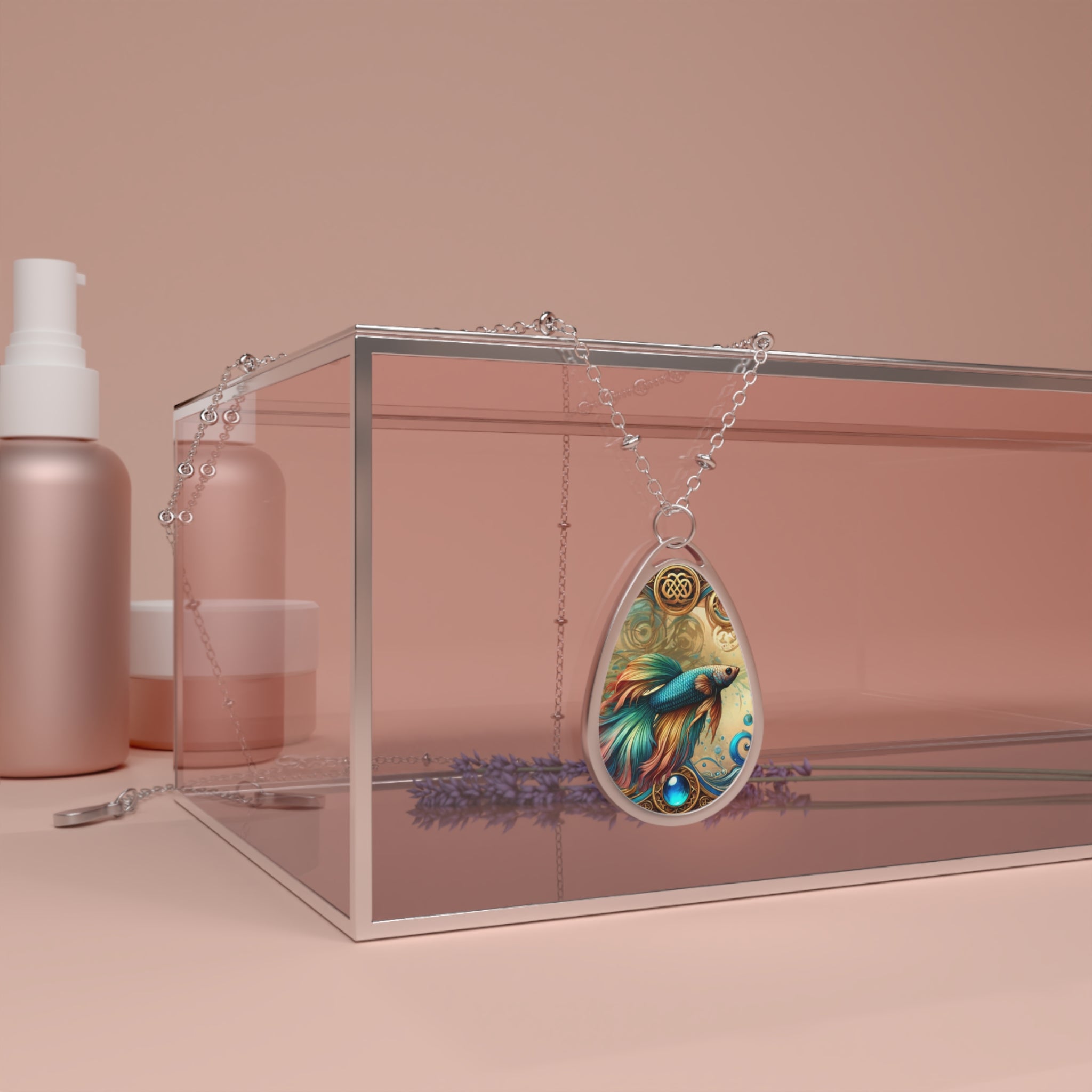 A close-up of a teardrop-shaped Betta Fish necklace displayed on a transparent box with a soft pink background. The necklace features a vibrant Betta Fish design in teal, orange, and gold, with swirling accents and a gemstone. Lavender sprigs and minimalist skincare bottles complete the aesthetic.