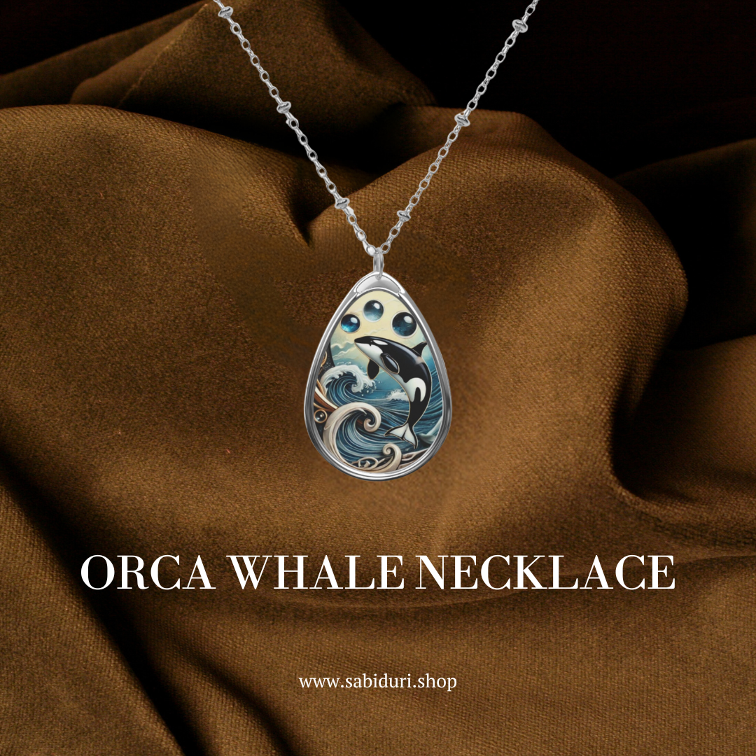 The orca whale pendant is showcased against a rich, brown fabric backdrop, highlighting its intricate design of the orca in stylized waves, topped with three blue gemstones. The text at the bottom reads "ORCA WHALE NECKLACE" and "www.sabiduri.shop.