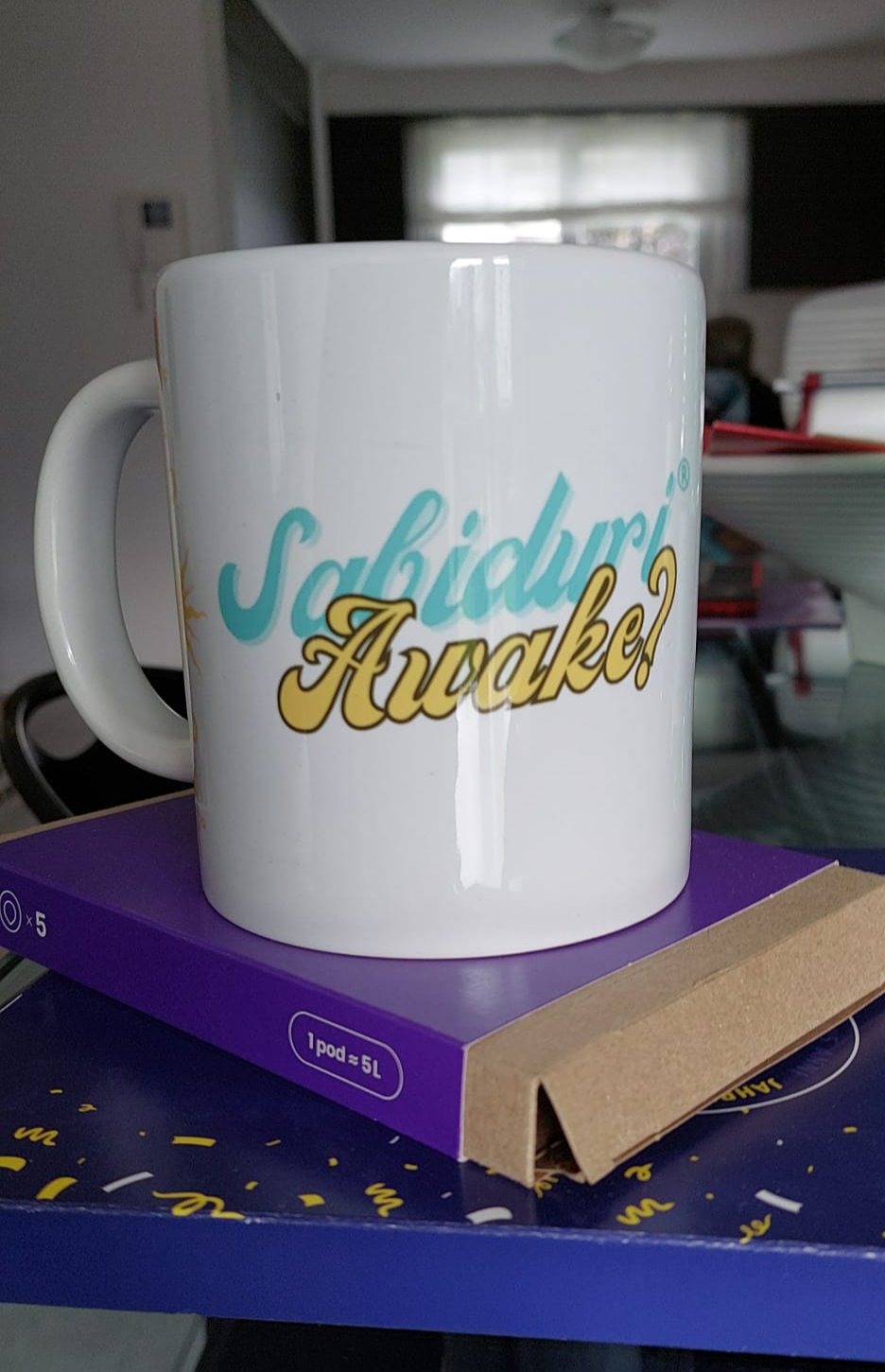 A close-up of a white ceramic mug with the phrase "Sabiduría Awake?" in bold letters, set against a casual indoor background with a glimpse of a kitchen and some clutter.