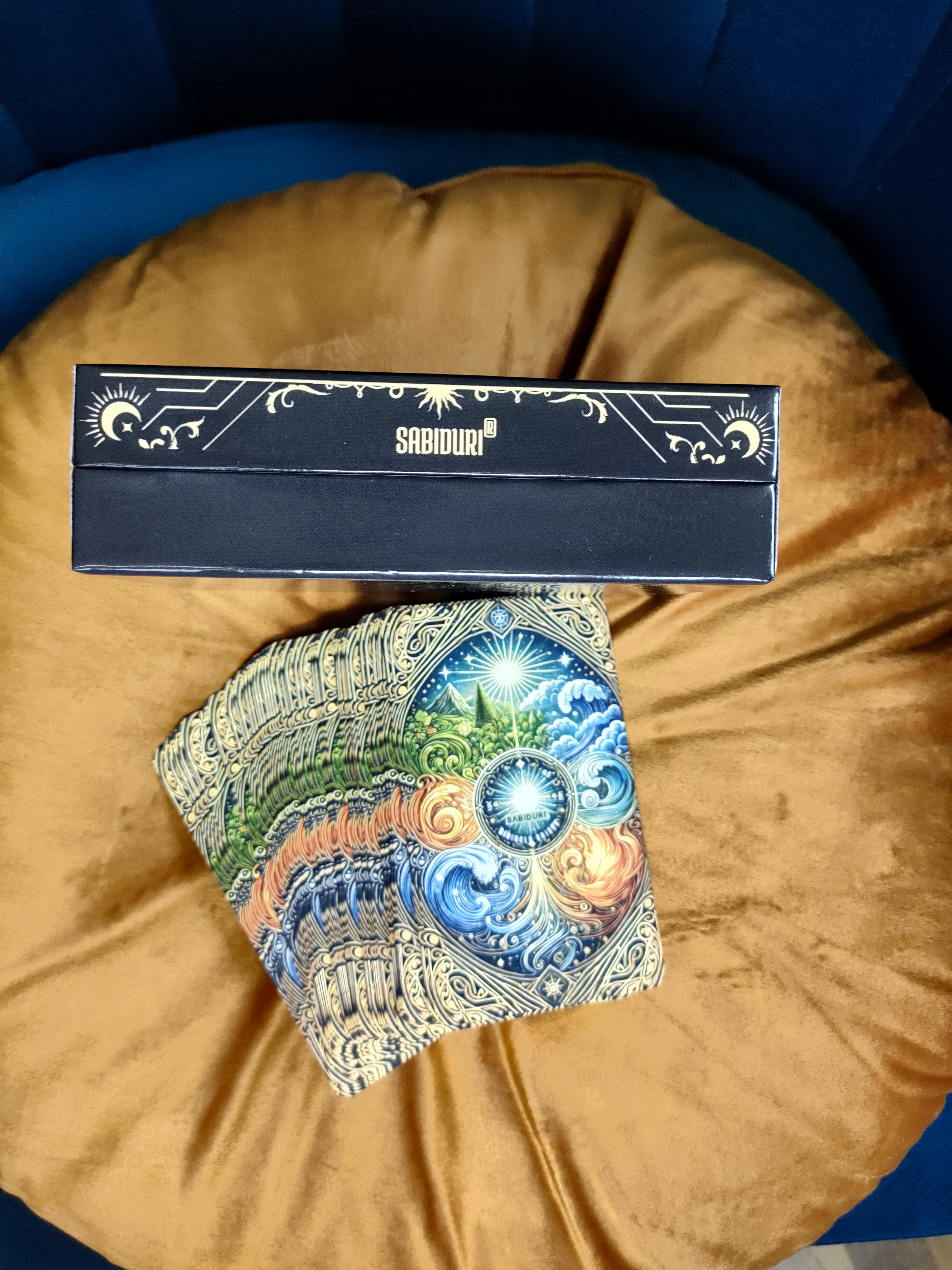 The side view of the Sabiduri Tarot deck box on the same mustard pillow, with "SABIDURIĀ®" written in gold on the side, surrounded by celestial designs, including a crescent moon and stars. Below the box, part of the tarot cards with the same swirling elemental design can be seen.