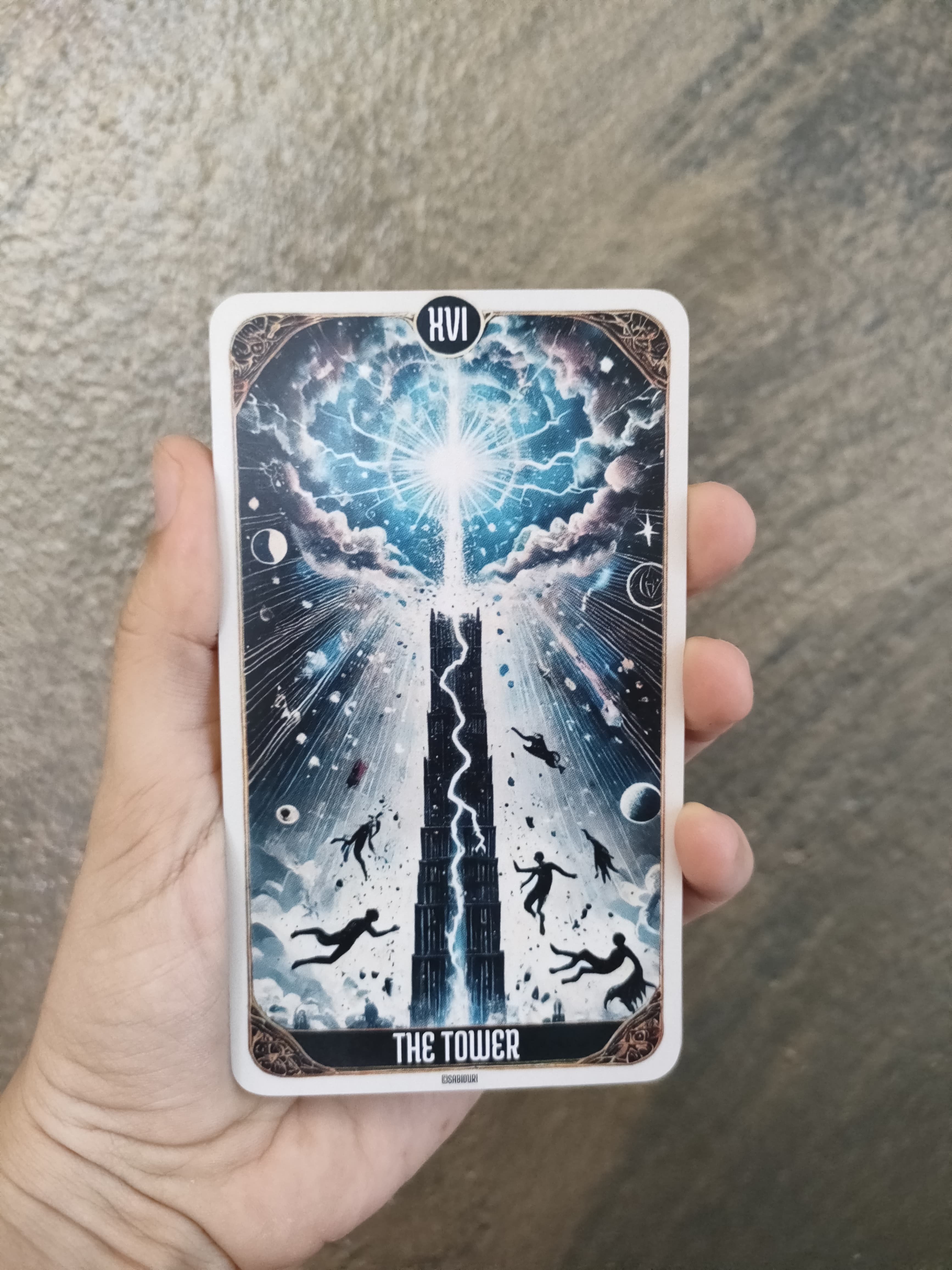 A close-up of "The Tower" tarot card from the Sabiduri deck, held in a hand. The card shows a towering structure being struck by lightning, with figures falling from it, symbolizing sudden change and upheaval.
