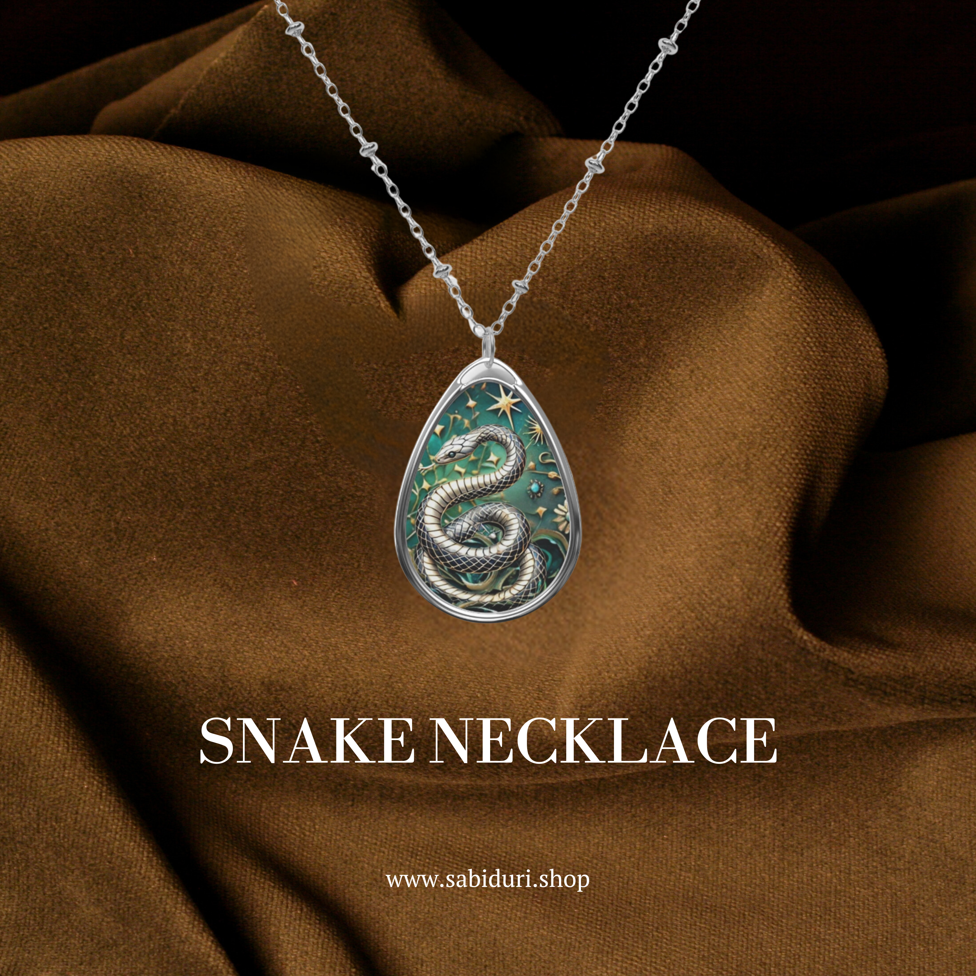 A snake pendant necklace with intricate details of a silver snake coiled against a green background adorned with stars. The necklace is set against a rich brown fabric backdrop with the text "Snake Necklace" and the website "www.sabiduri.shop" displayed below.