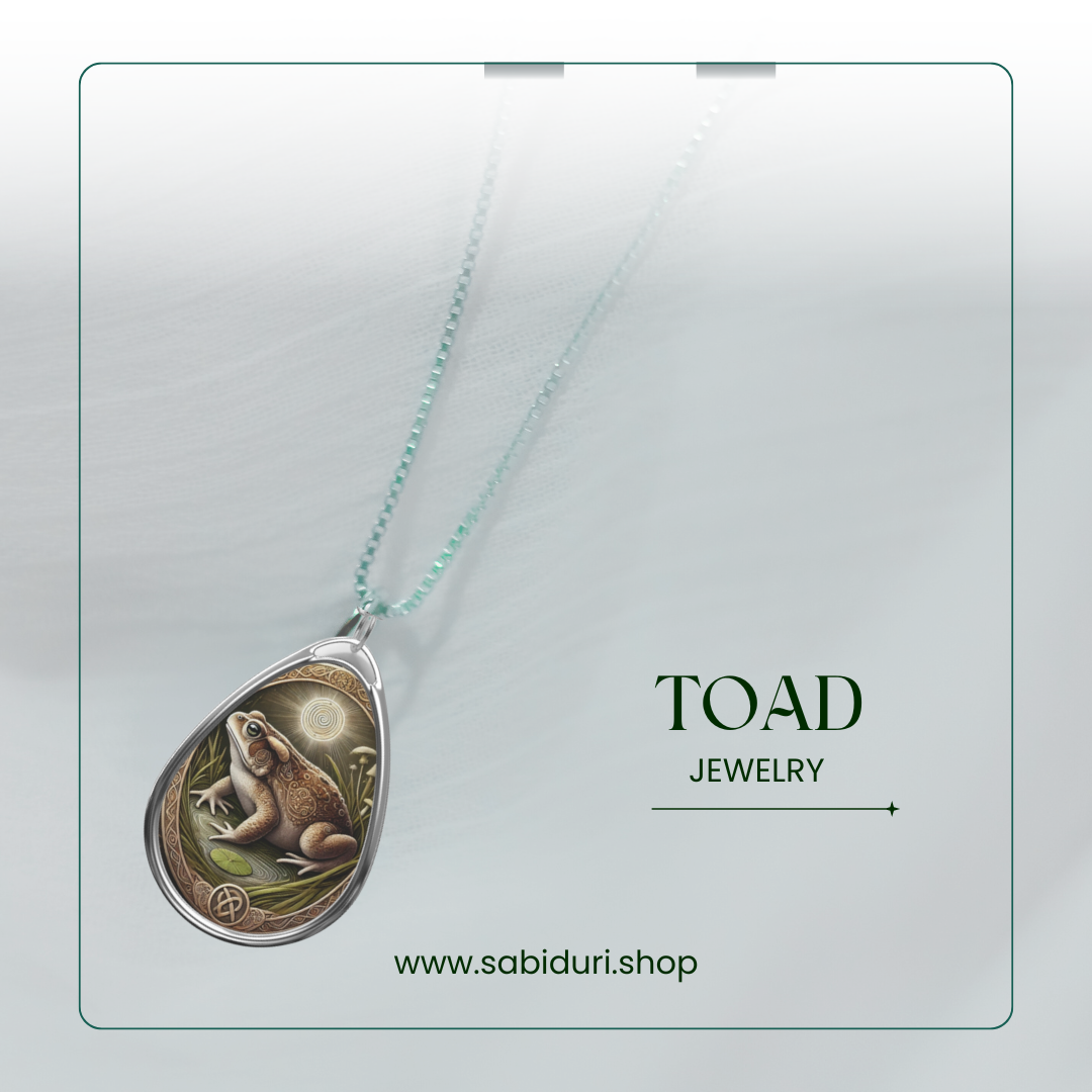 A toad pendant necklace displayed on a soft white background. The pendant showcases a toad surrounded by foliage and mystical spiral symbols, representing resilience and adaptability. The text reads "TOAD Jewelry" along with the website "www.sabiduri.shop.